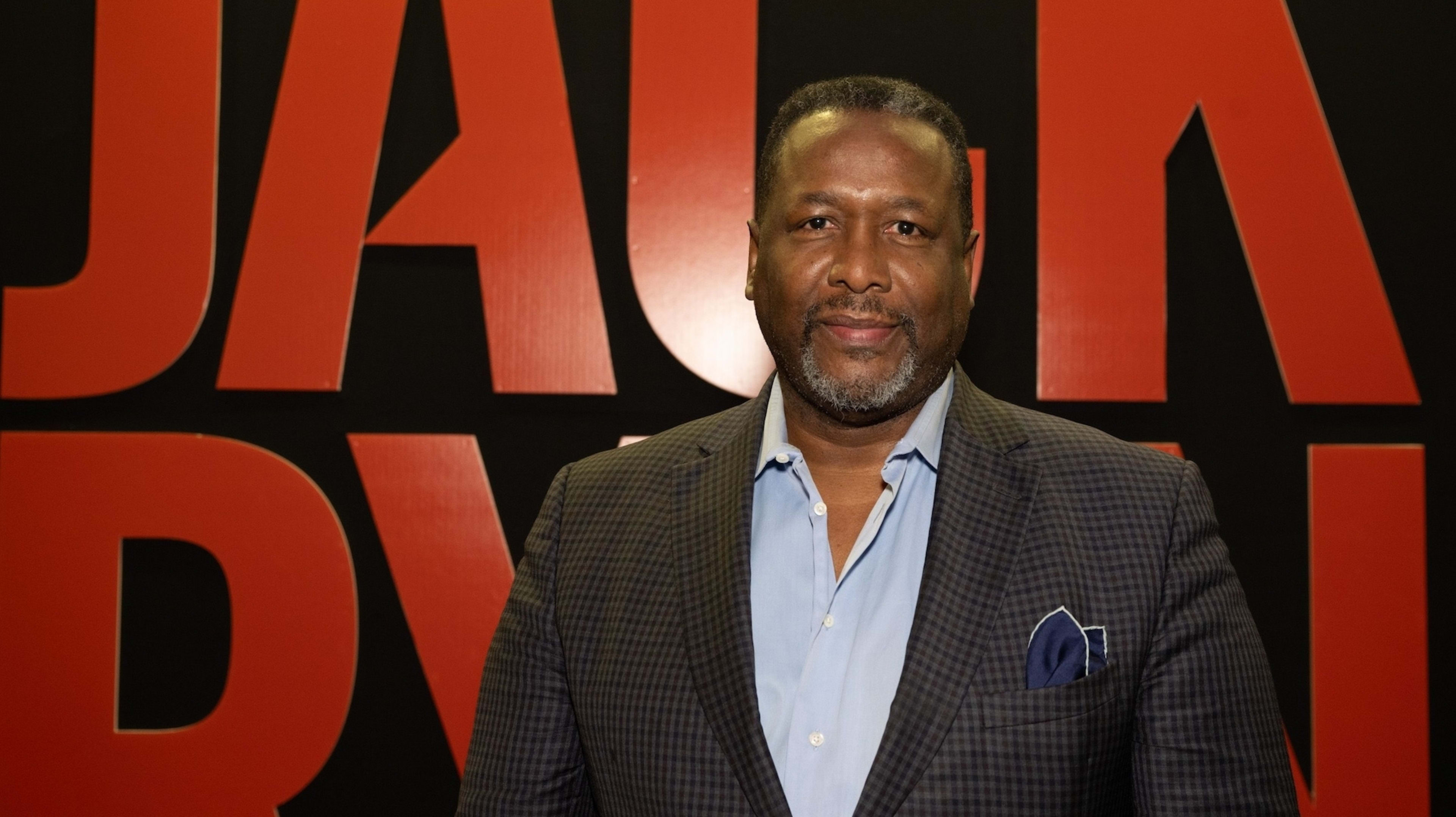 A man in a suit jacket and blue shirt stands in front of a backdrop with large, bold text