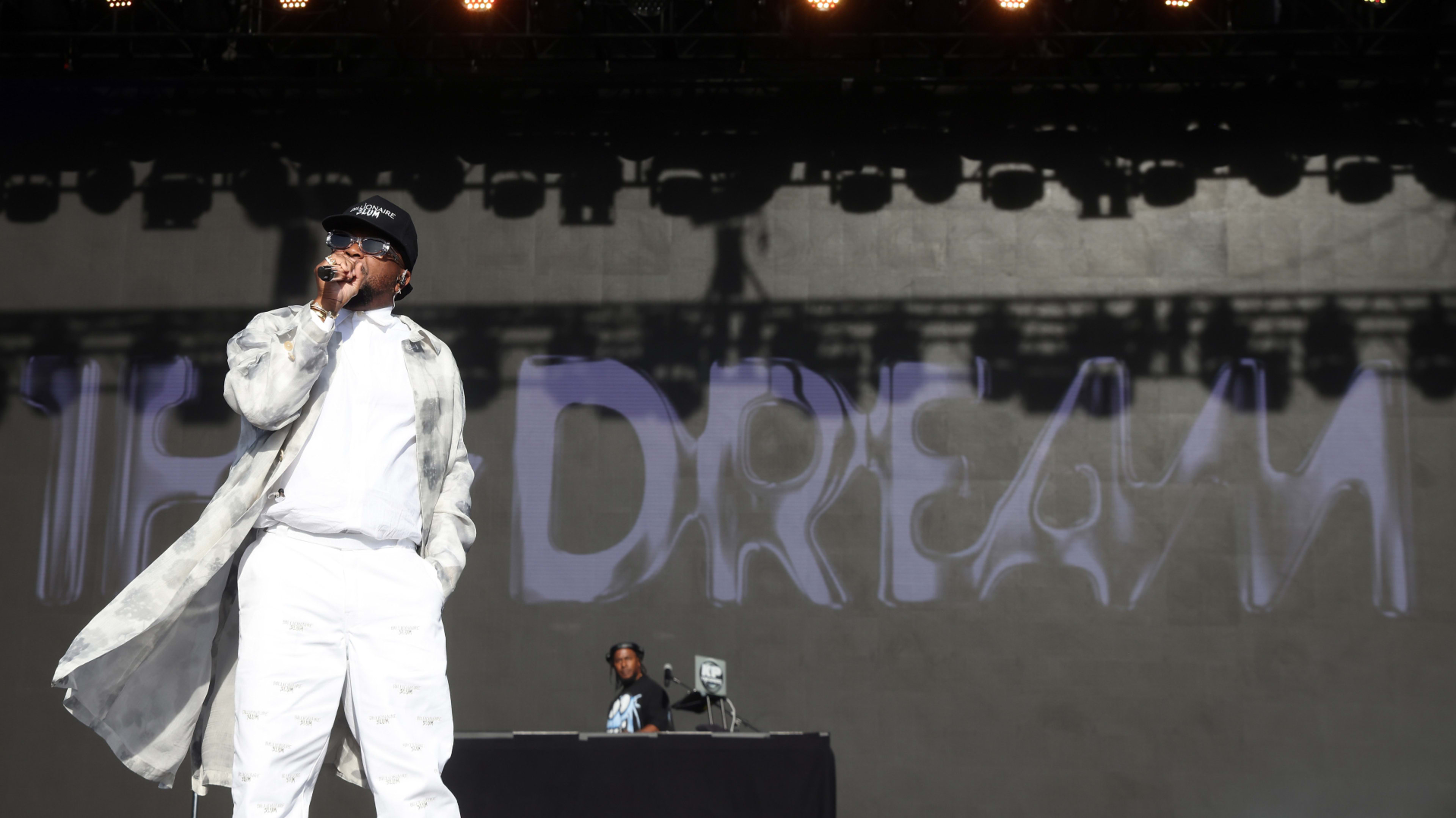 Dream performs on stage in a white outfit and sunglasses. Large text reading "The Dream" is displayed on the screen behind him. A DJ is in the background