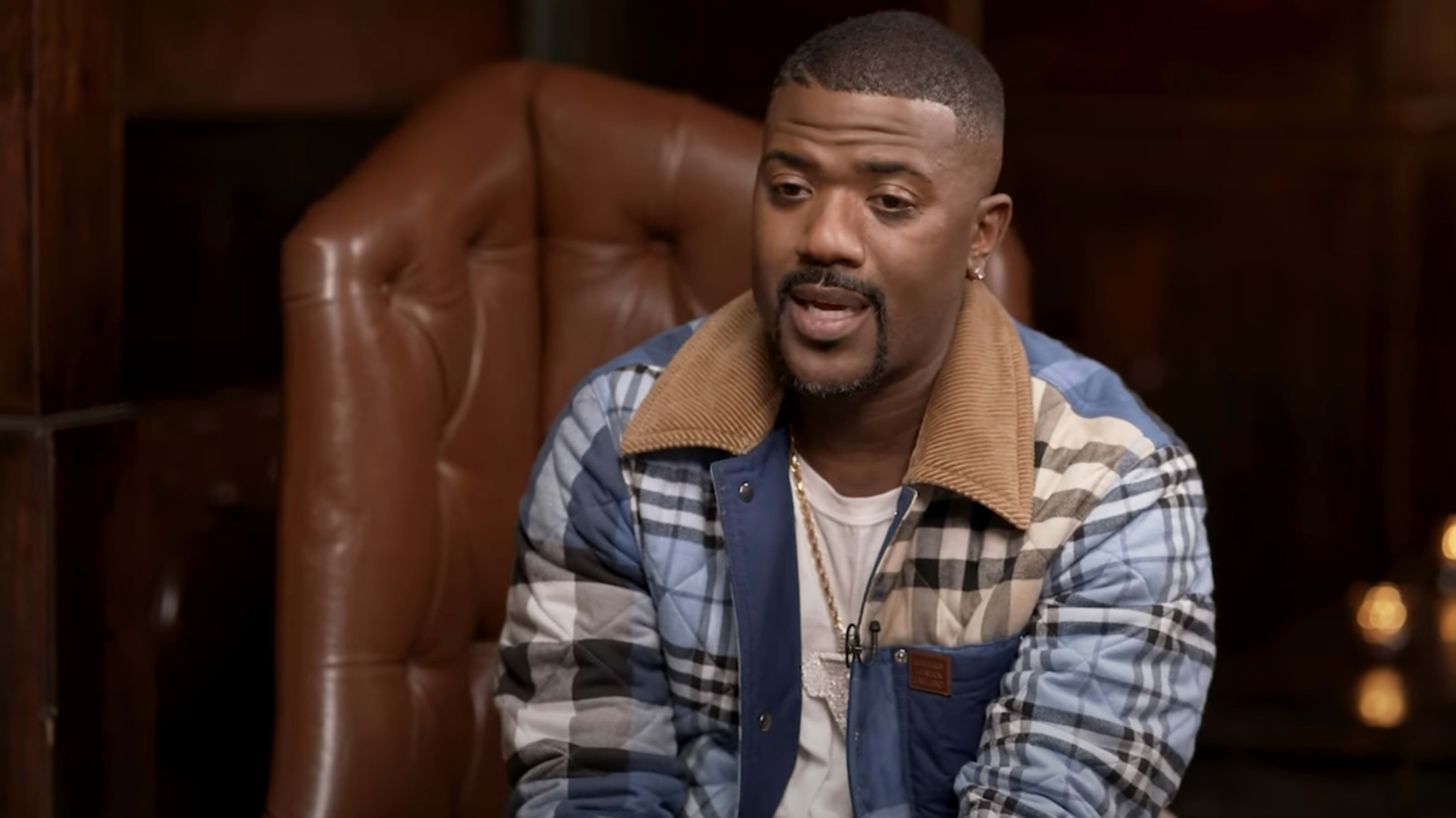 Ray J sits in a leather armchair wearing a quilted jacket with a plaid pattern and a chain necklace