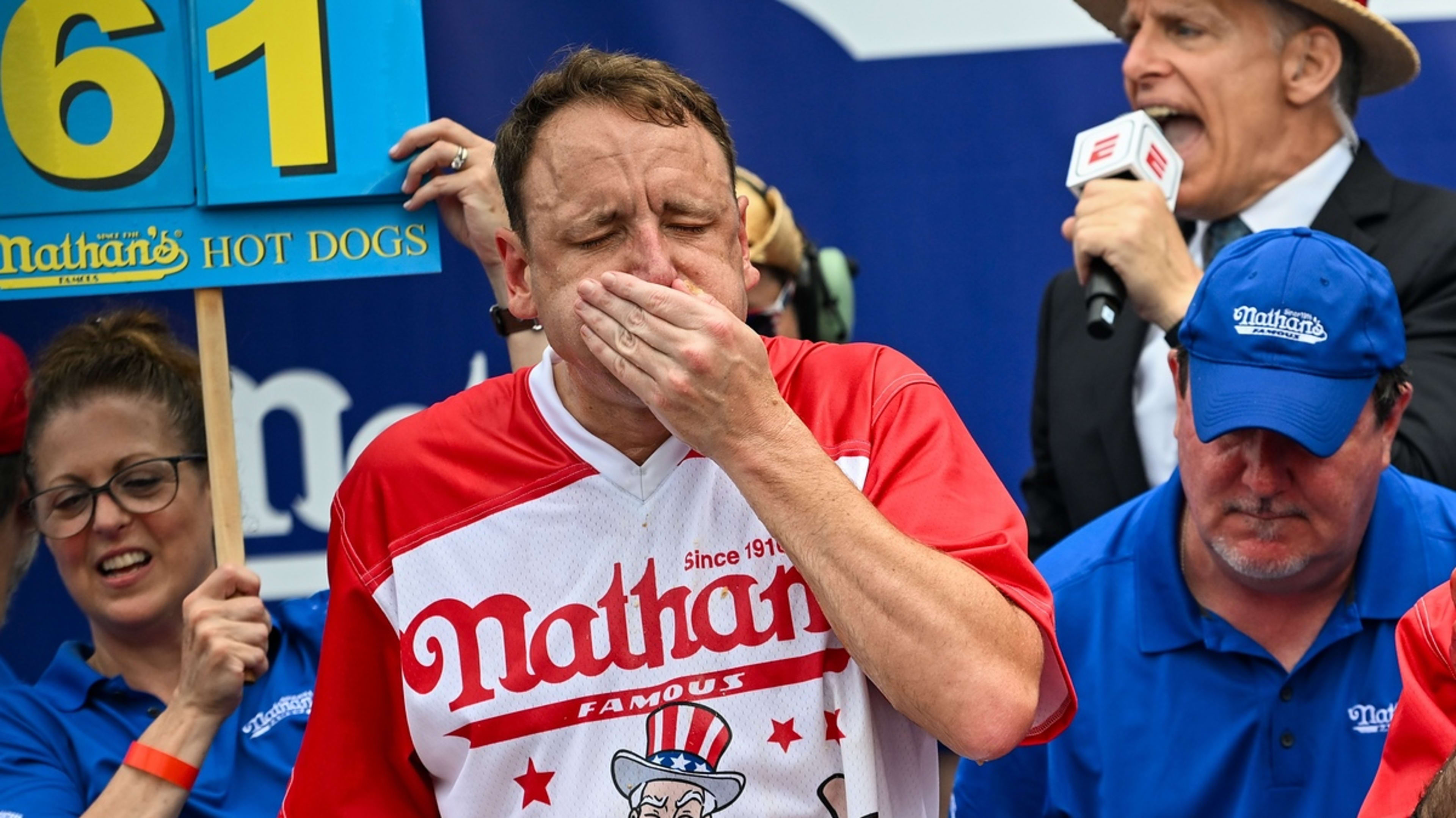 Joey Chestnut, in a Nathan's shirt, competes in a hot dog eating contest, covering his mouth. Announcer and assistants are visible in the background. Sign shows 61