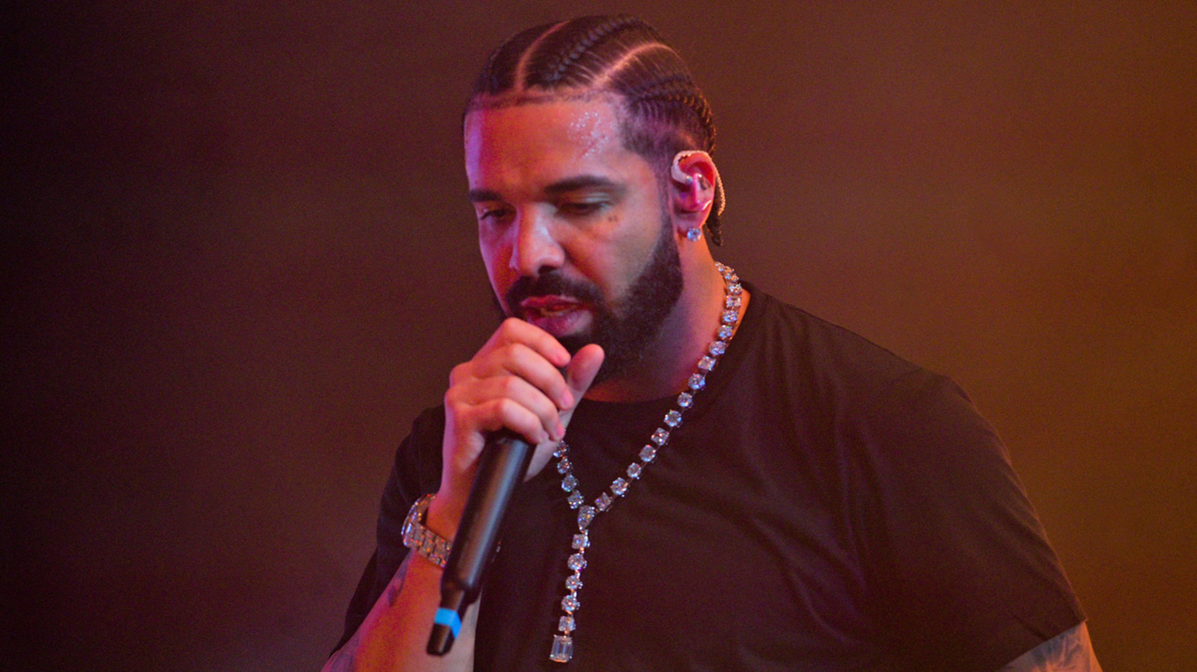 Drake performs on stage, holding a microphone and wearing a black shirt and a beaded necklace