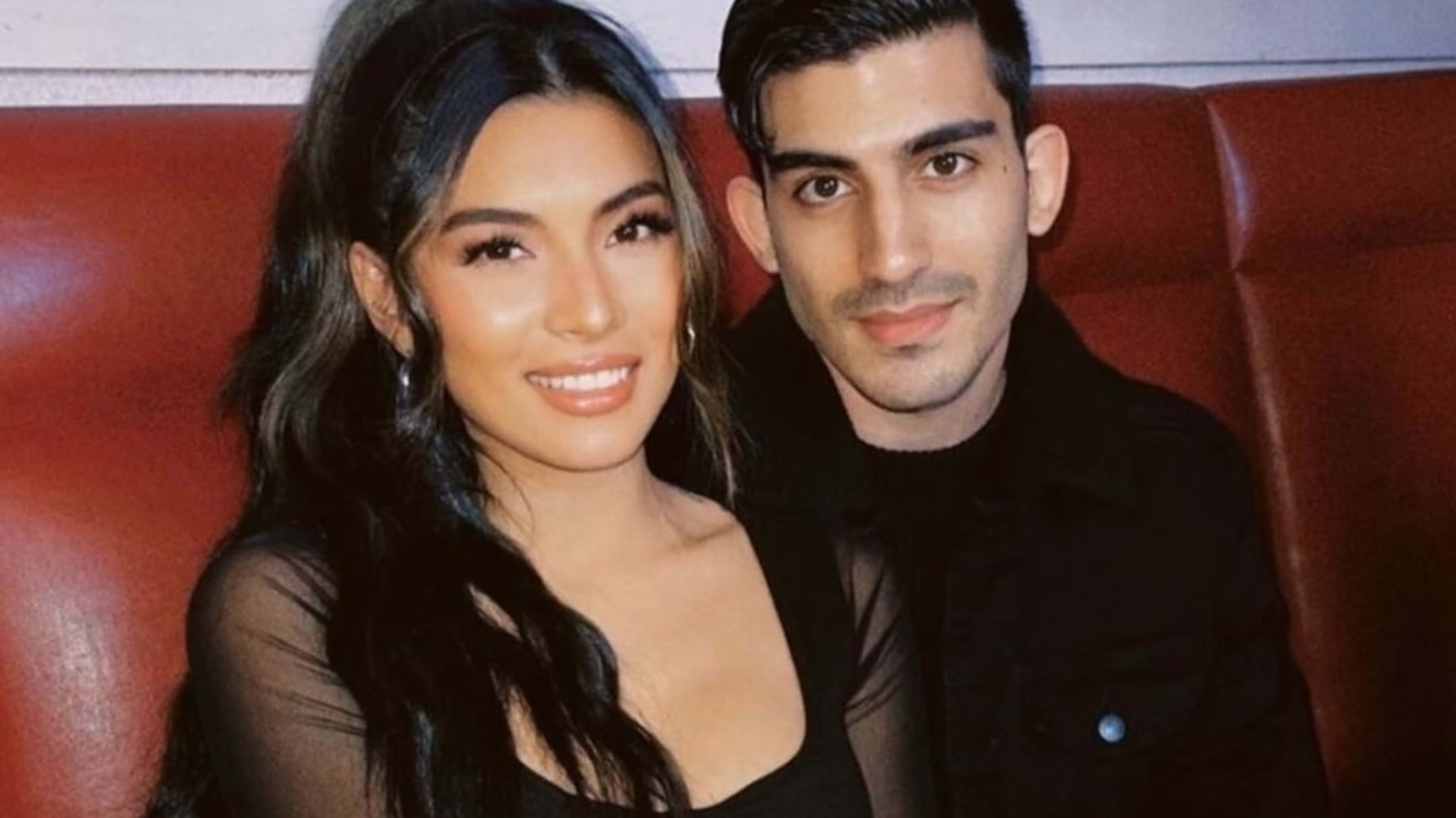 A man and woman, both smiling, sit close together in a booth. The woman wears a sheer black top, and the man is dressed in a dark shirt. Their names are not known