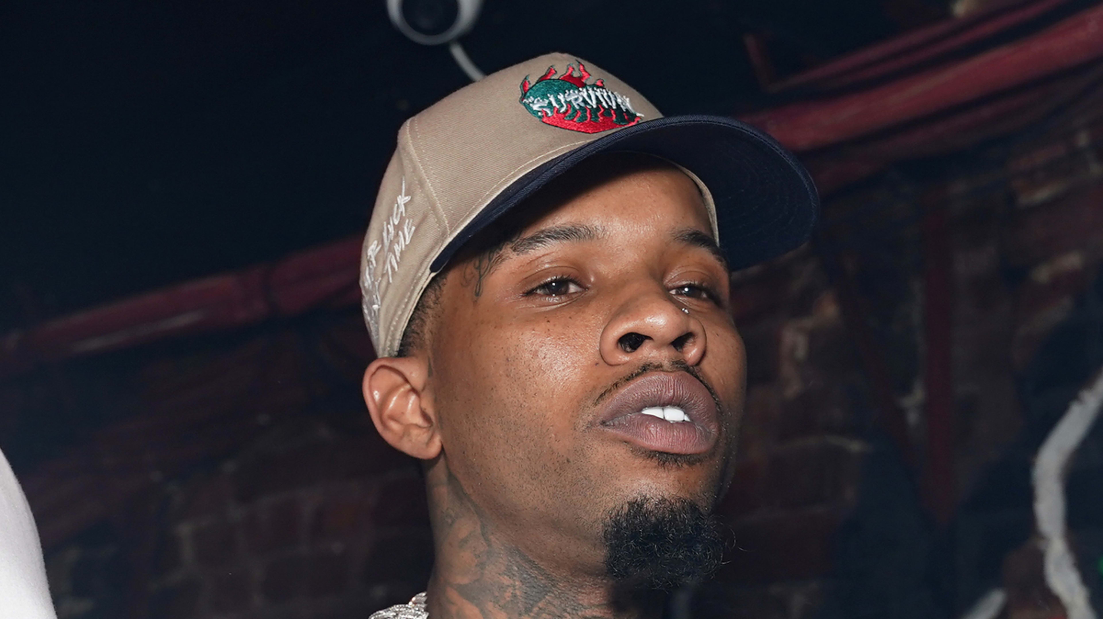 Tory Lanez wearing a white T-shirt, beige cap with a red logo, and layered chain necklaces, stands in an indoor setting