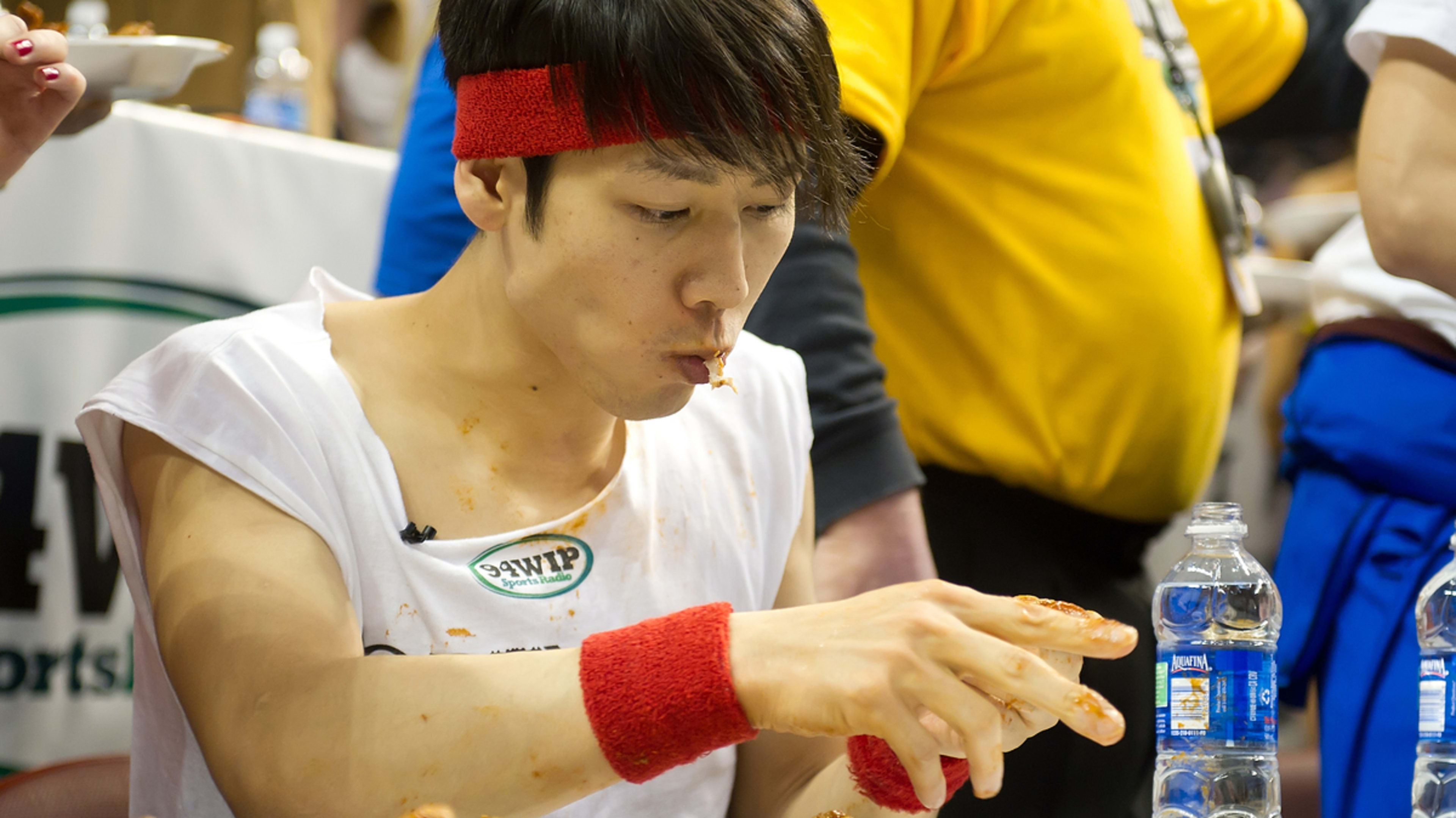 A man wearing a white tank top and red sweatbands is eating a large quantity of chicken wings in what appears to be a competitive eating contest. Other people are in the background