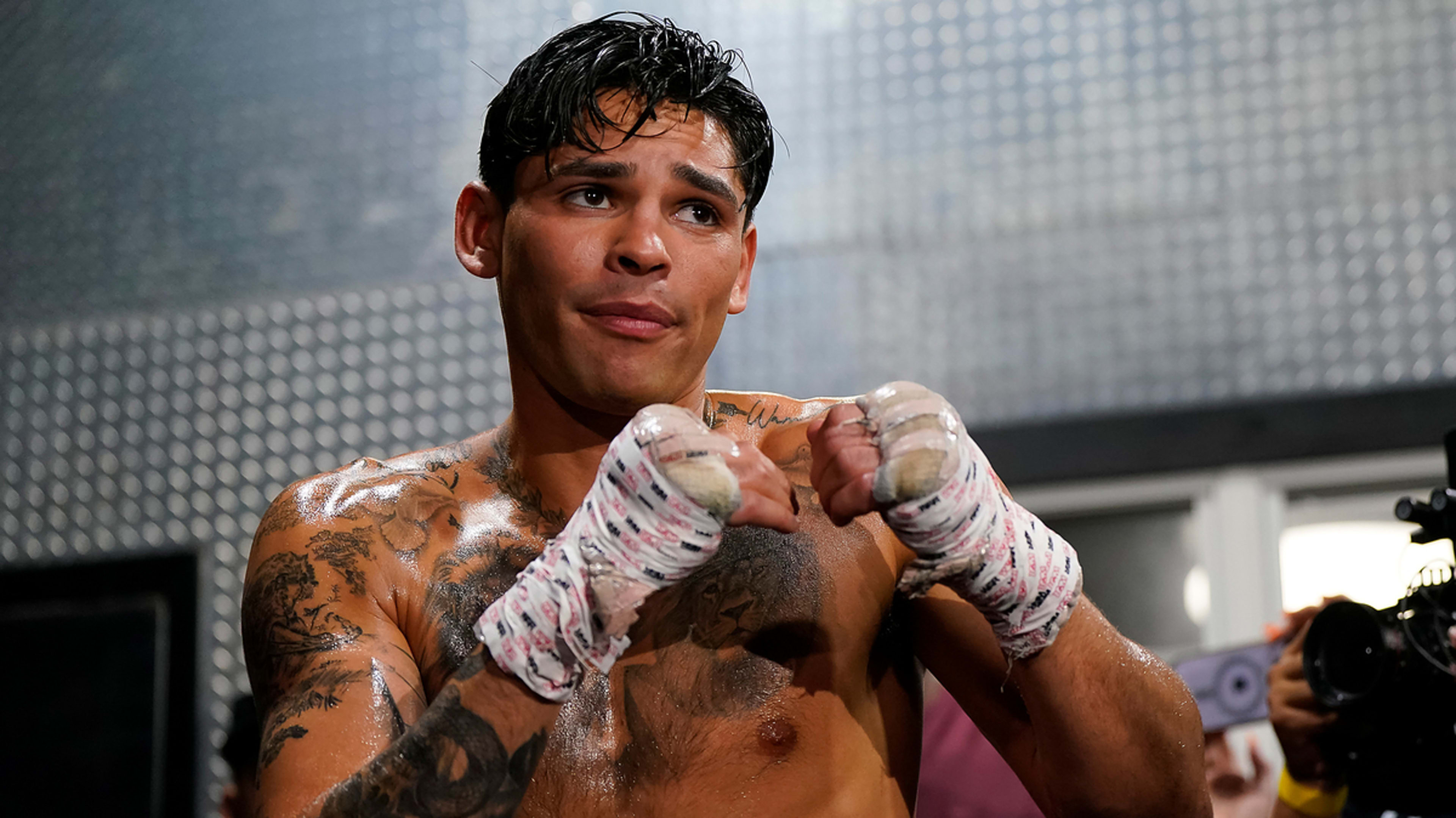 Ryan Garcia, with boxing gloves and hand wraps, stands in a boxing stance, shirtless, displaying tattoos on chest and arms, during a training session