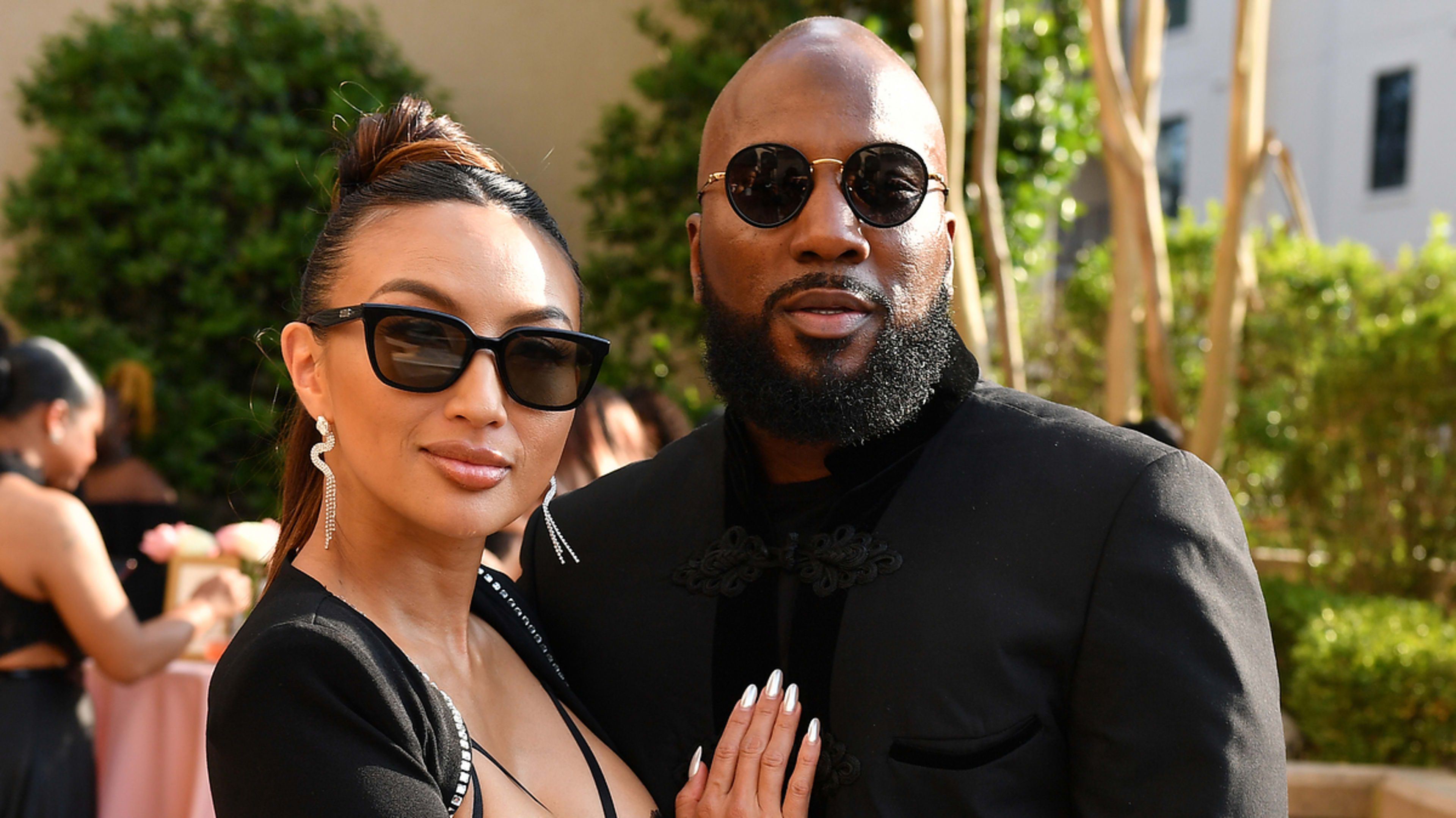 Jeannie Mai and Jeezy pose together outdoors; Jeannie wears a stylish black dress with a slit, and Jeezy sports a black jacket with round sunglasses