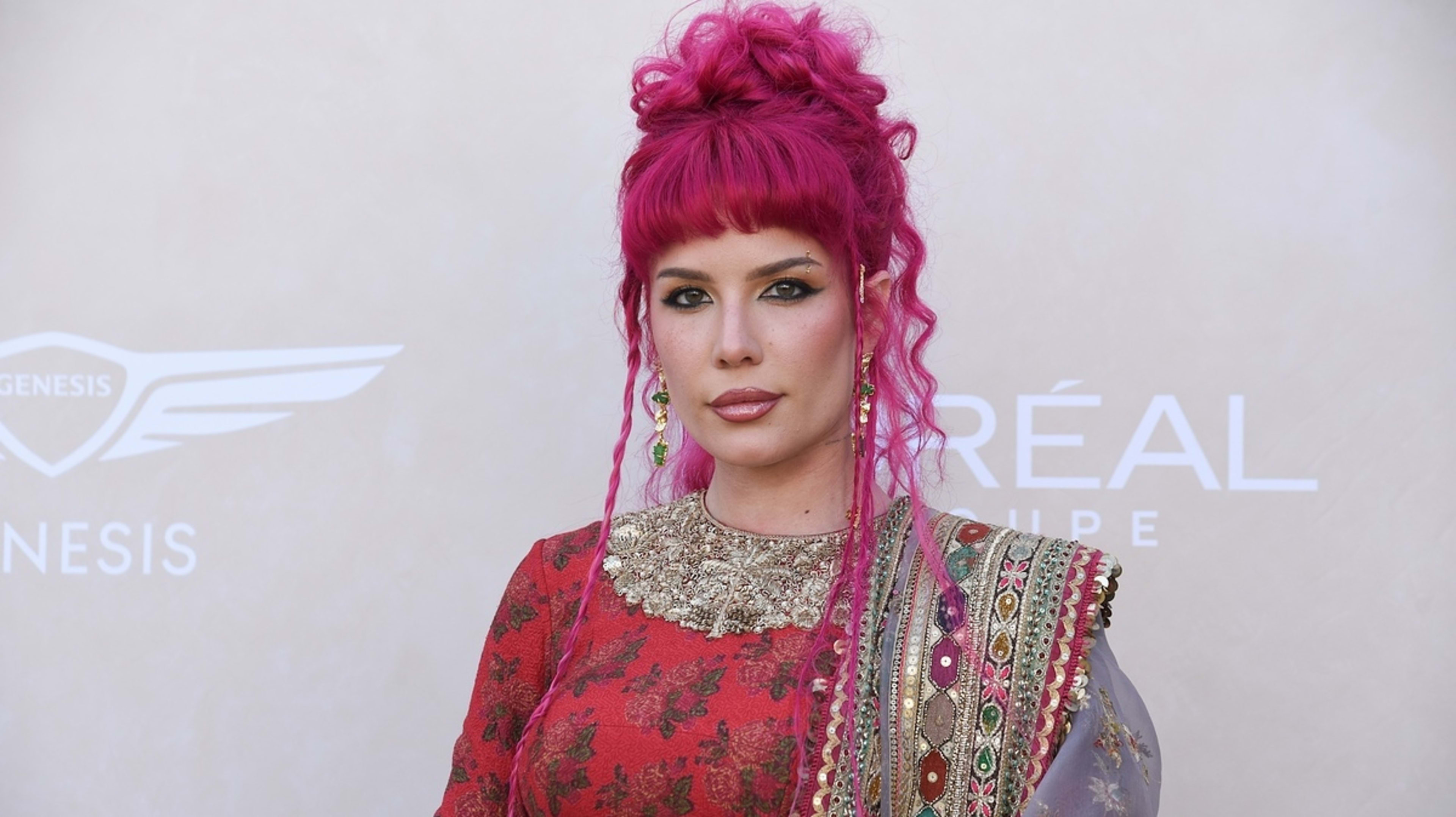 Halsey stands in front of a backdrop with "Genesis" and "L'Oreal Paris" logos, wearing an ornate, floral-patterned dress with intricate embroidered details and accessories