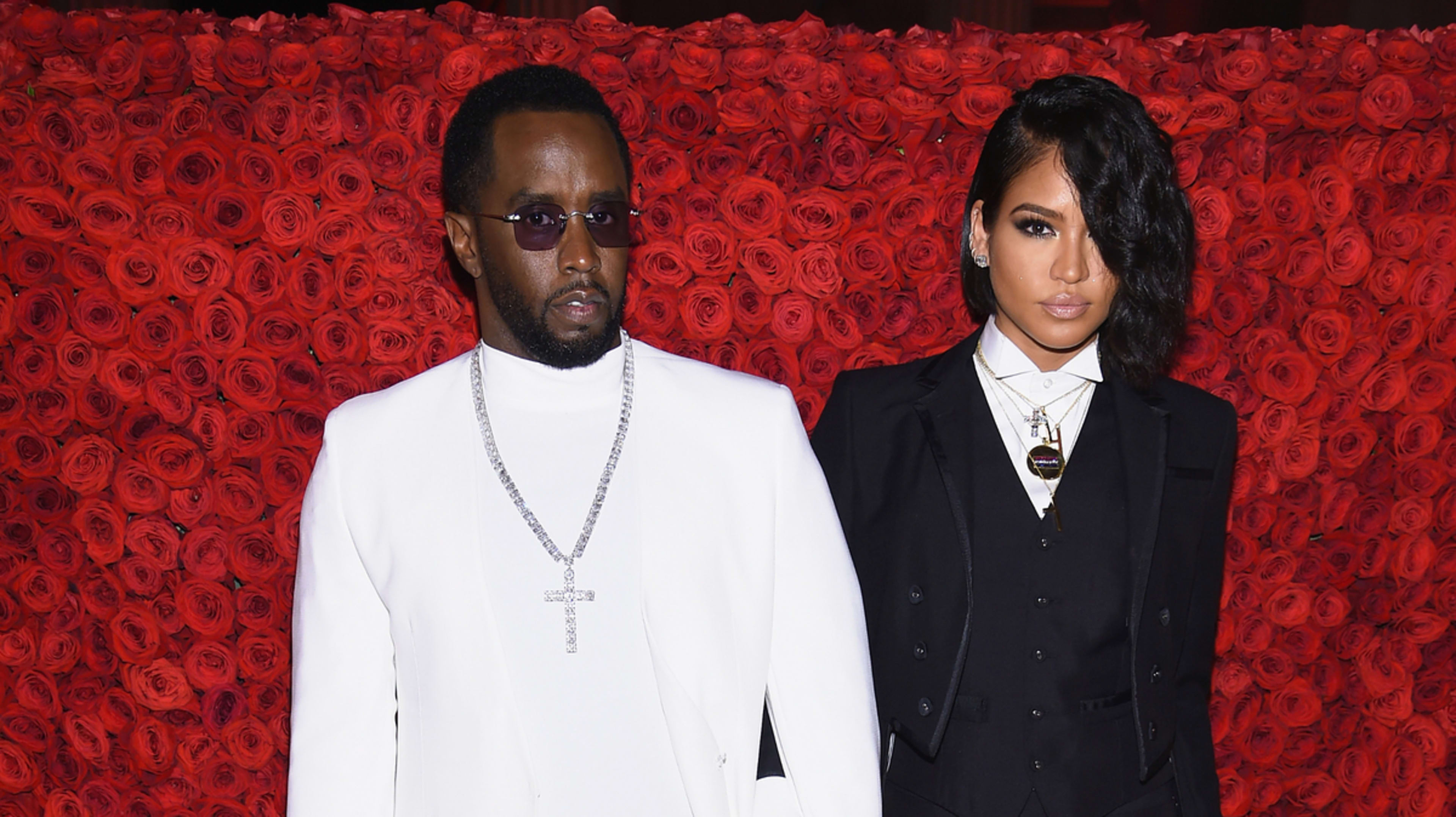 Sean “Diddy” Combs and Cassie Ventura pose together, with Diddy in a white suit and Cassie in a black suit at a red-carpet event, against a backdrop of red roses