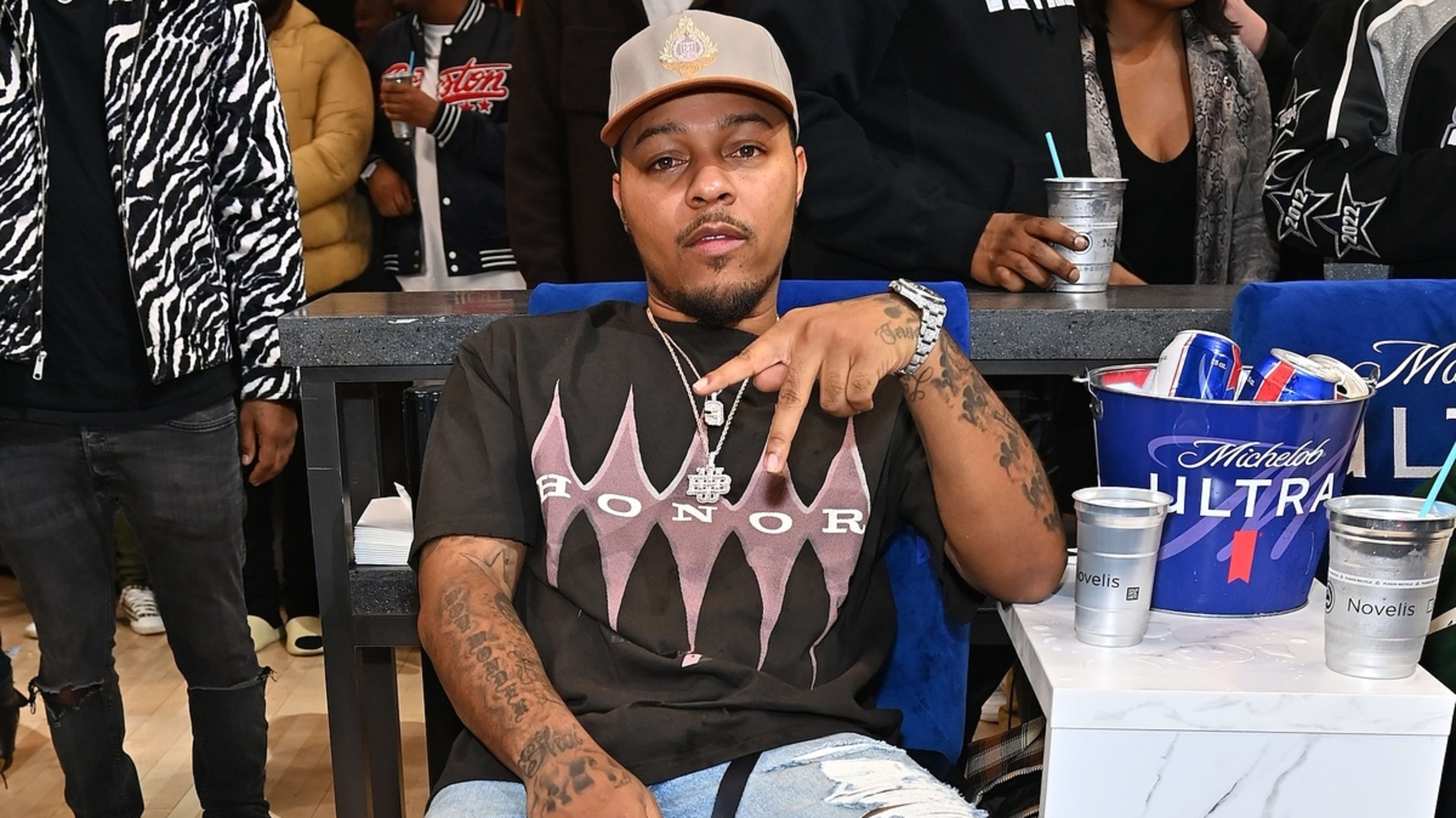 Bow Wow sits casually at an event, wearing a cap and a black Honor T-shirt with ripped jeans, making a hand gesture. Several people are in the background