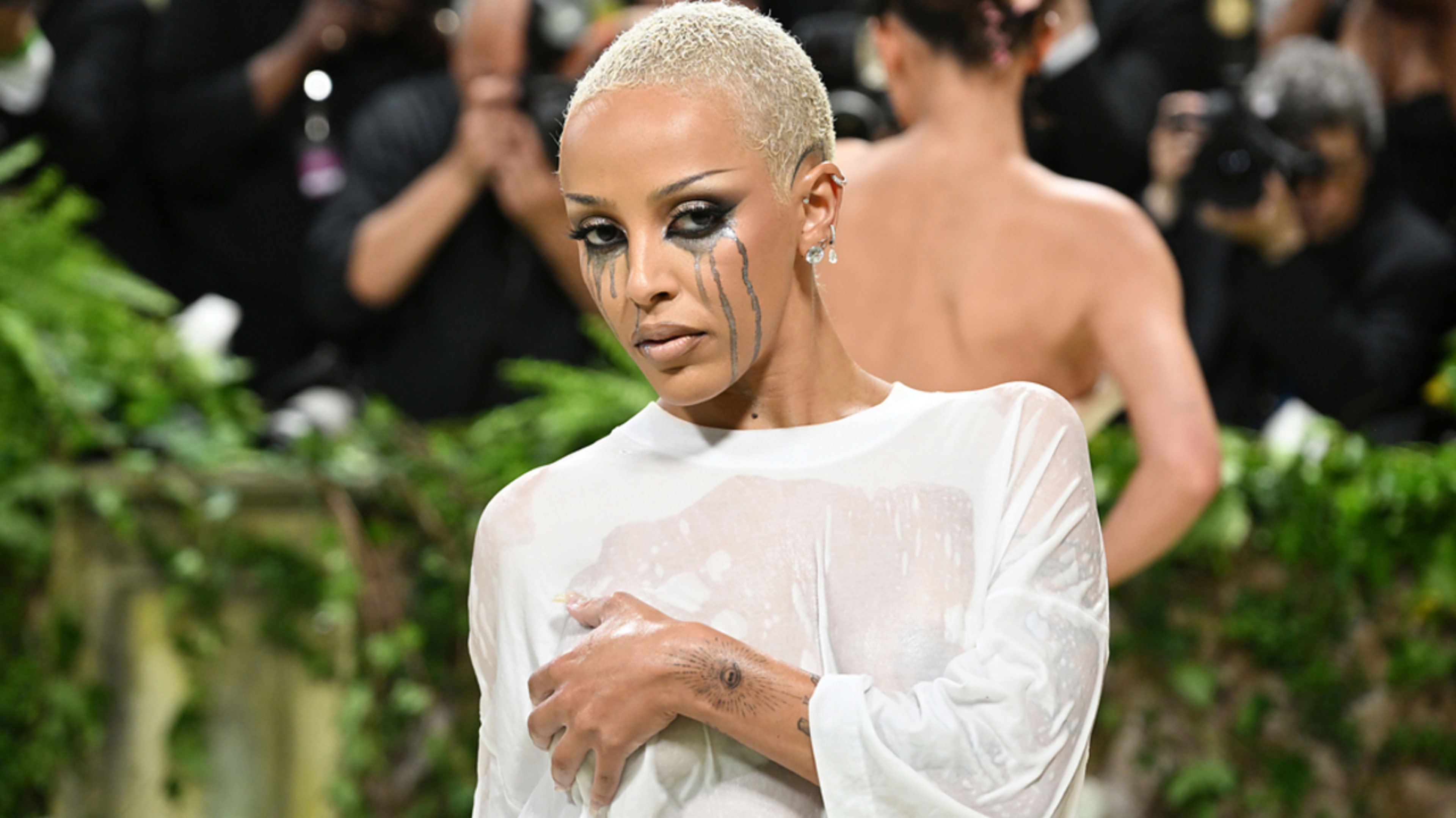 Doja Cat wears silver earrings and a sheer outfit on the Met Gala red carpet, posing with dramatic black face makeup resembling streaking tears