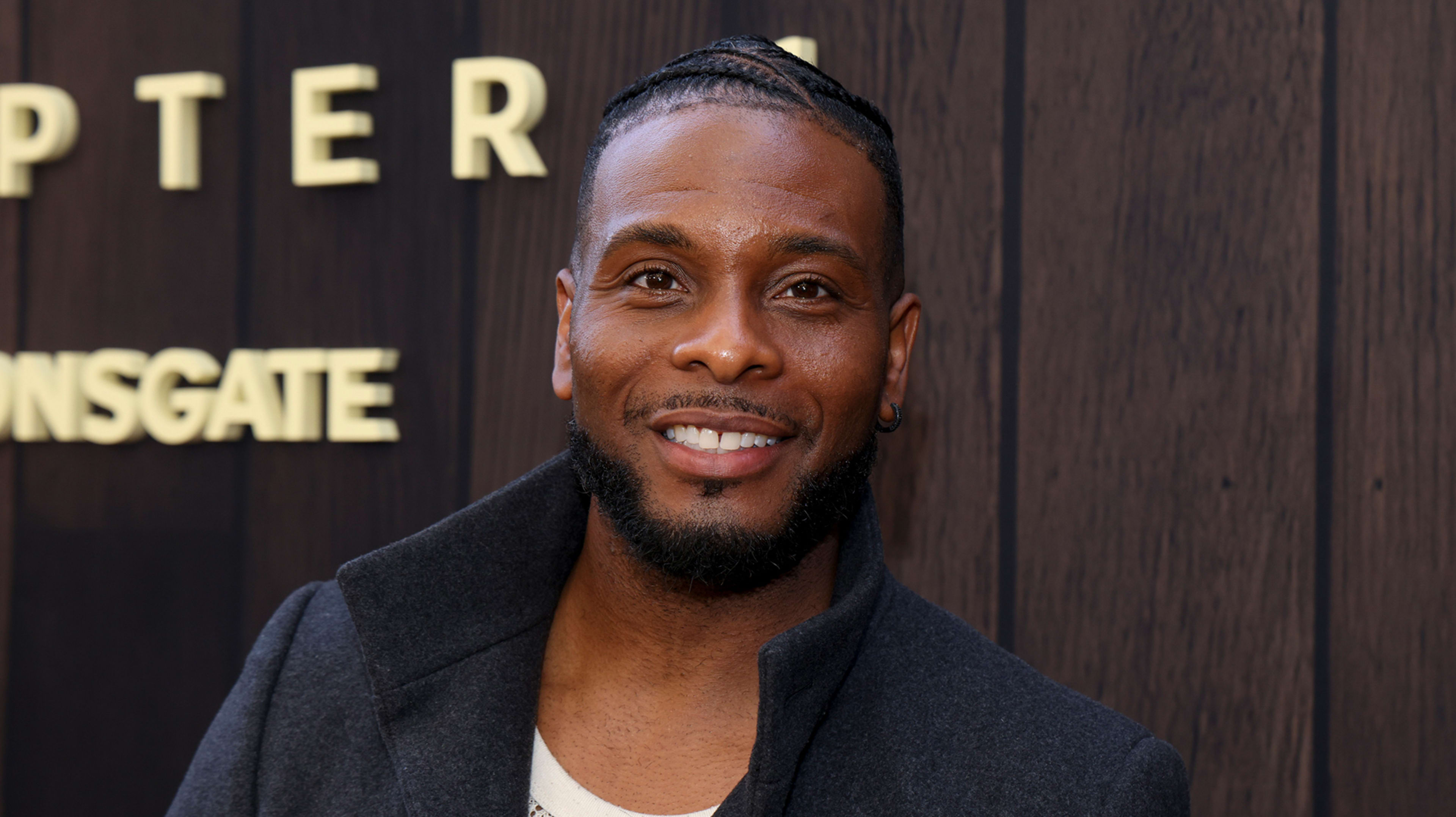 Kel Mitchell at a pop culture event, wearing a casual jacket over a t-shirt, standing in front of a wooden backdrop with movie studio branding