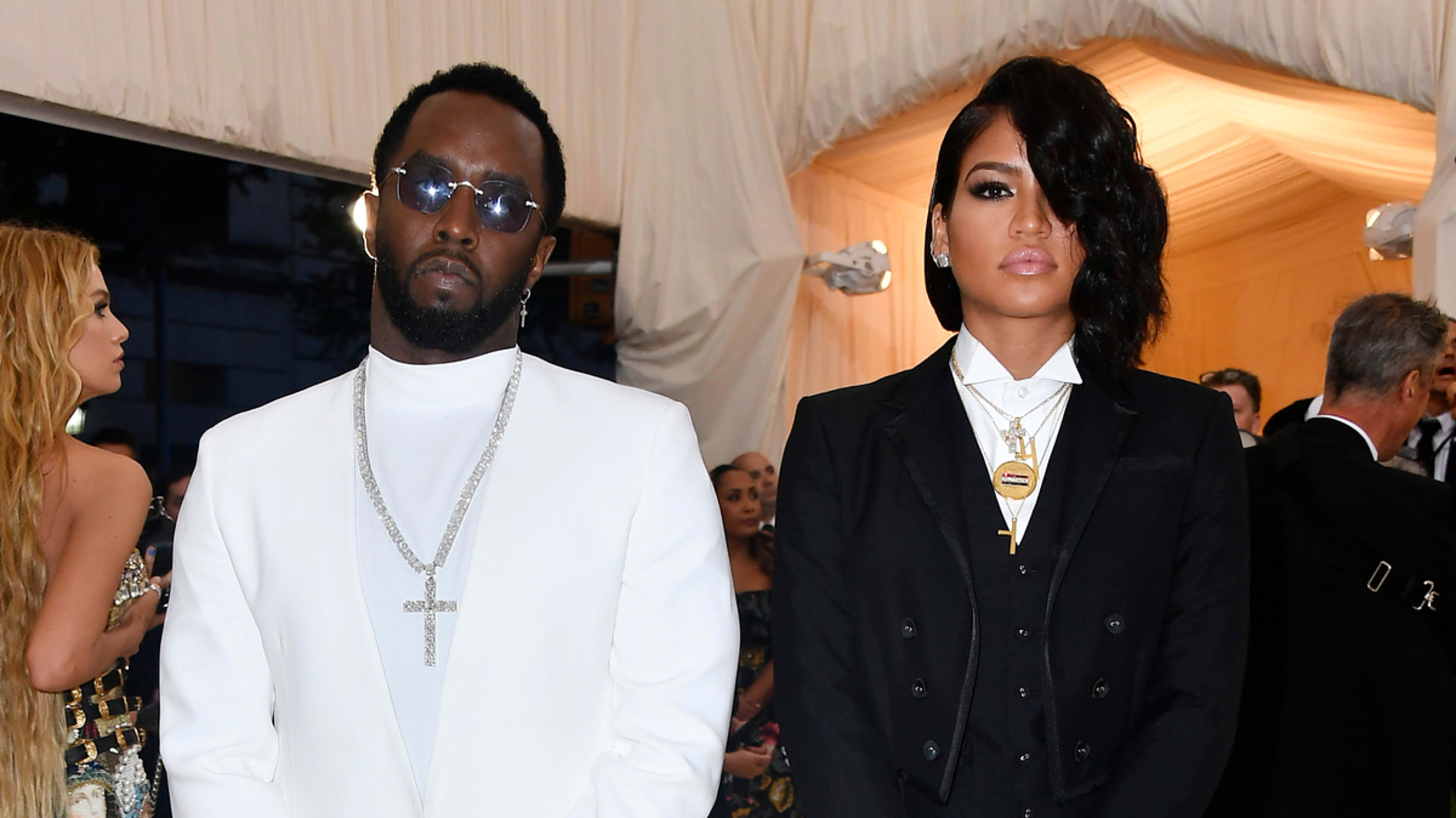 Sean Combs in a white suit and Cassie Ventura in a black suit attend a formal event with other attendees in the background