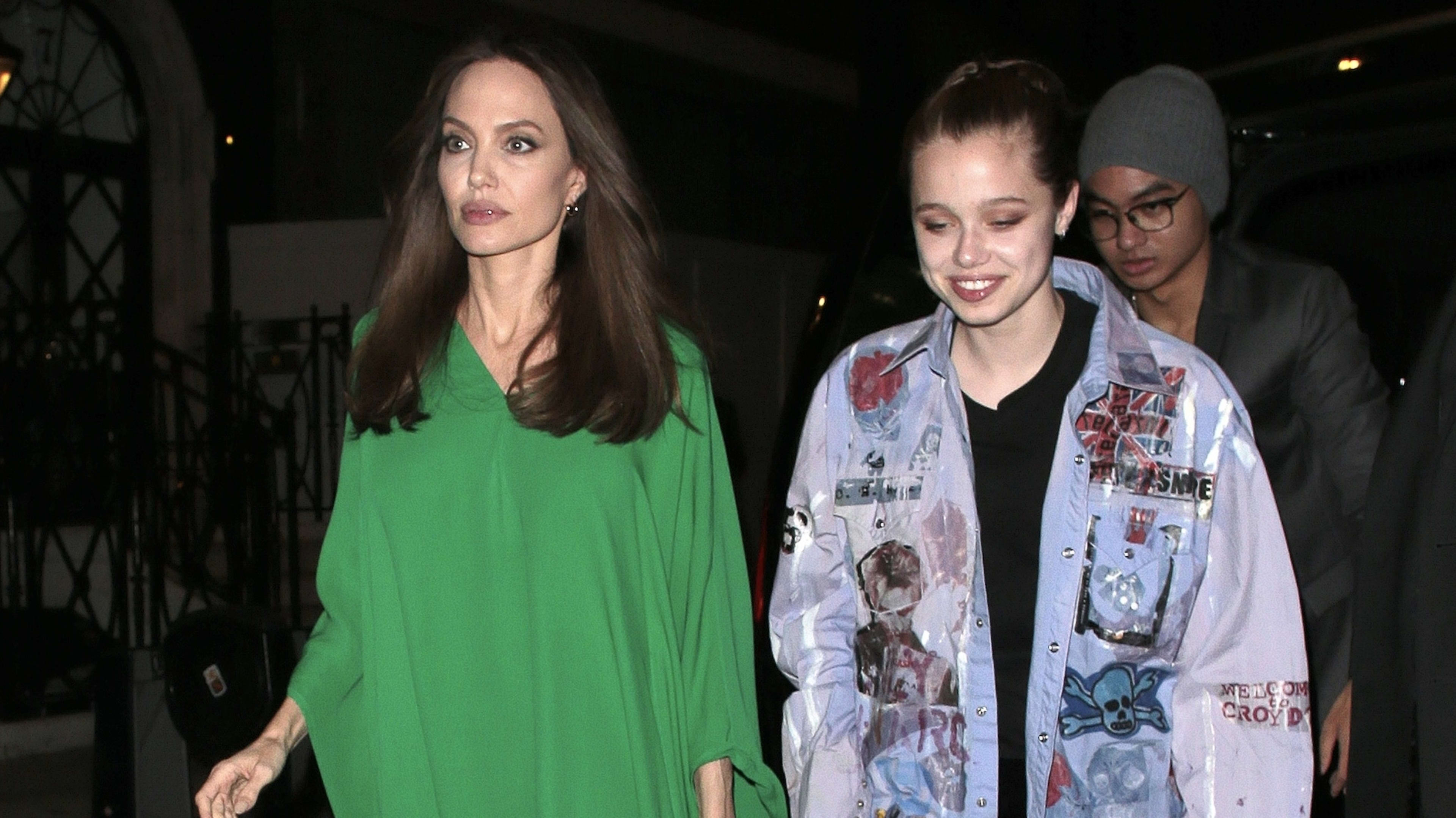 Angelina Jolie in a flowing green dress walks beside Shiloh Jolie-Pitt, who wears a painted jacket. A third person in a beanie is in the background