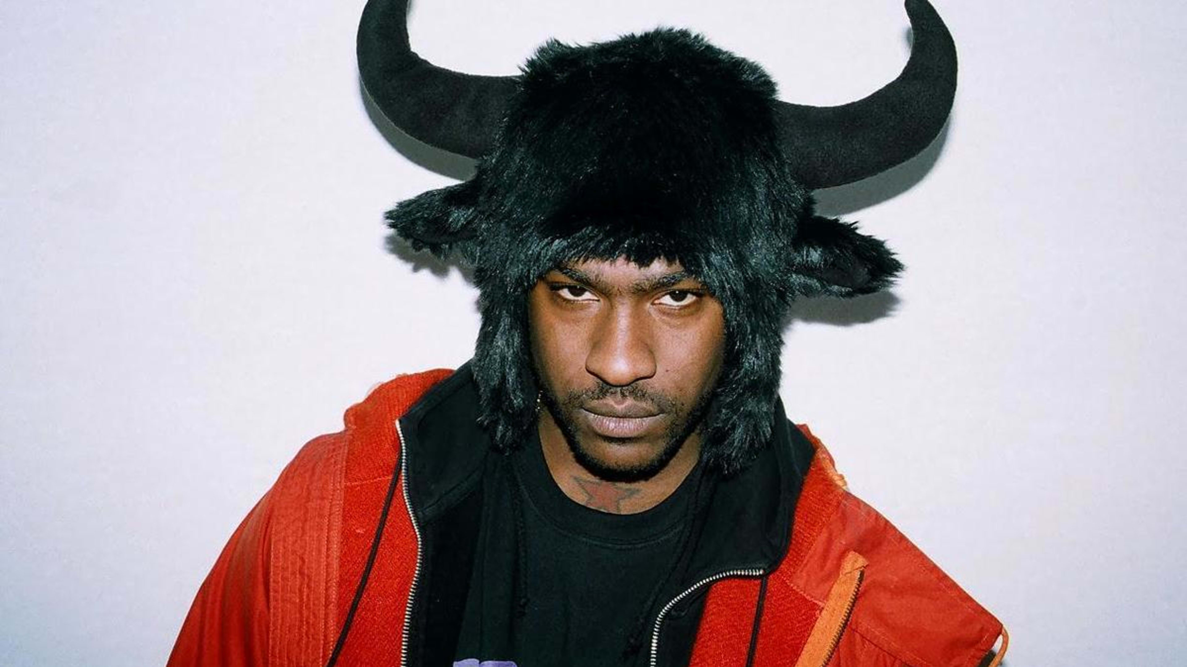 Skepta wearing a black furry hat with bull horns, a black graphic T-shirt, and an orange jacket, poses against a plain background