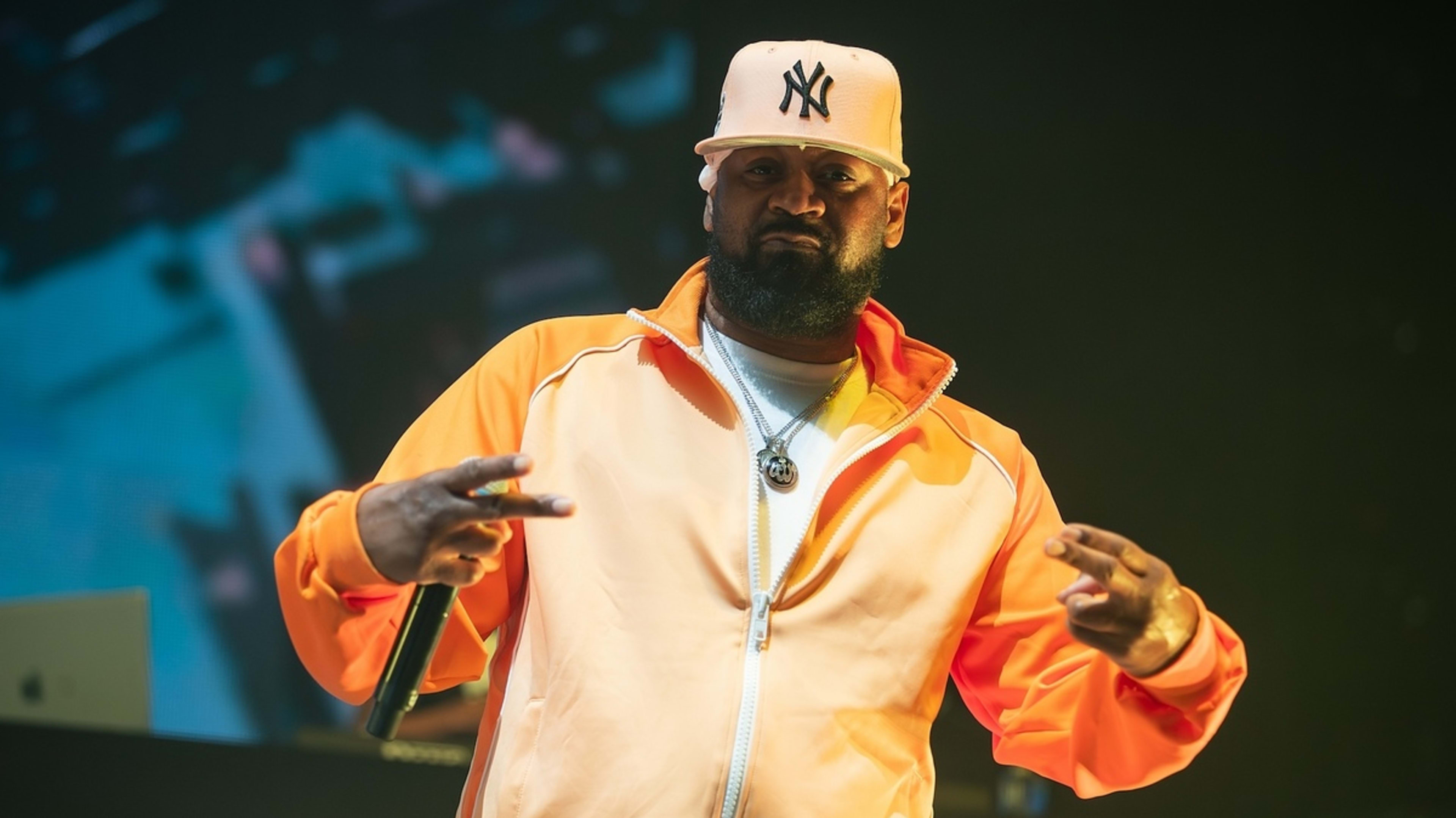 Music artist on stage wearing a baseball cap and an orange jacket, performing with a microphone stand in the background