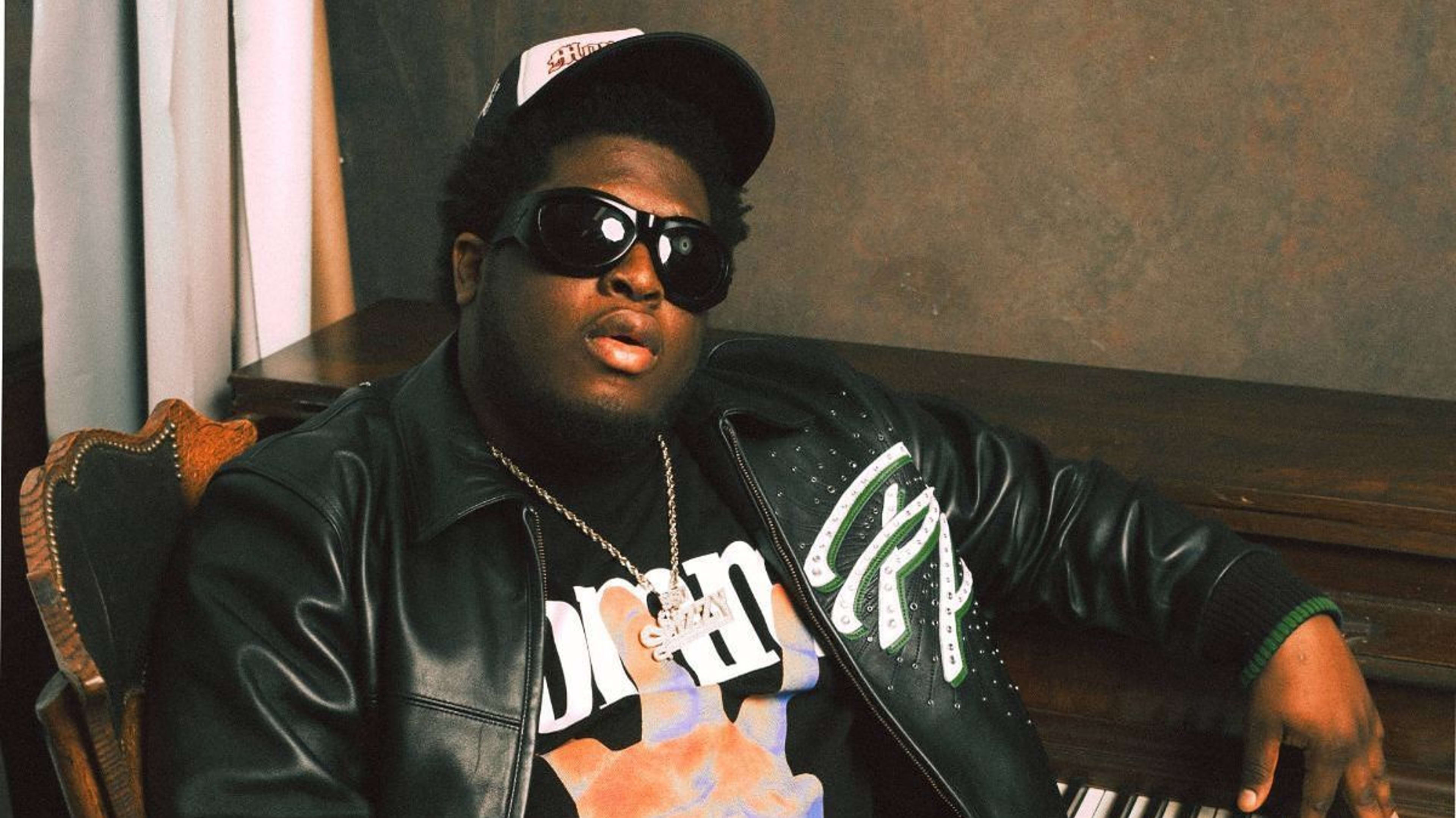 Musician in a leather jacket and sunglasses sits beside a piano, exuding a relaxed vibe