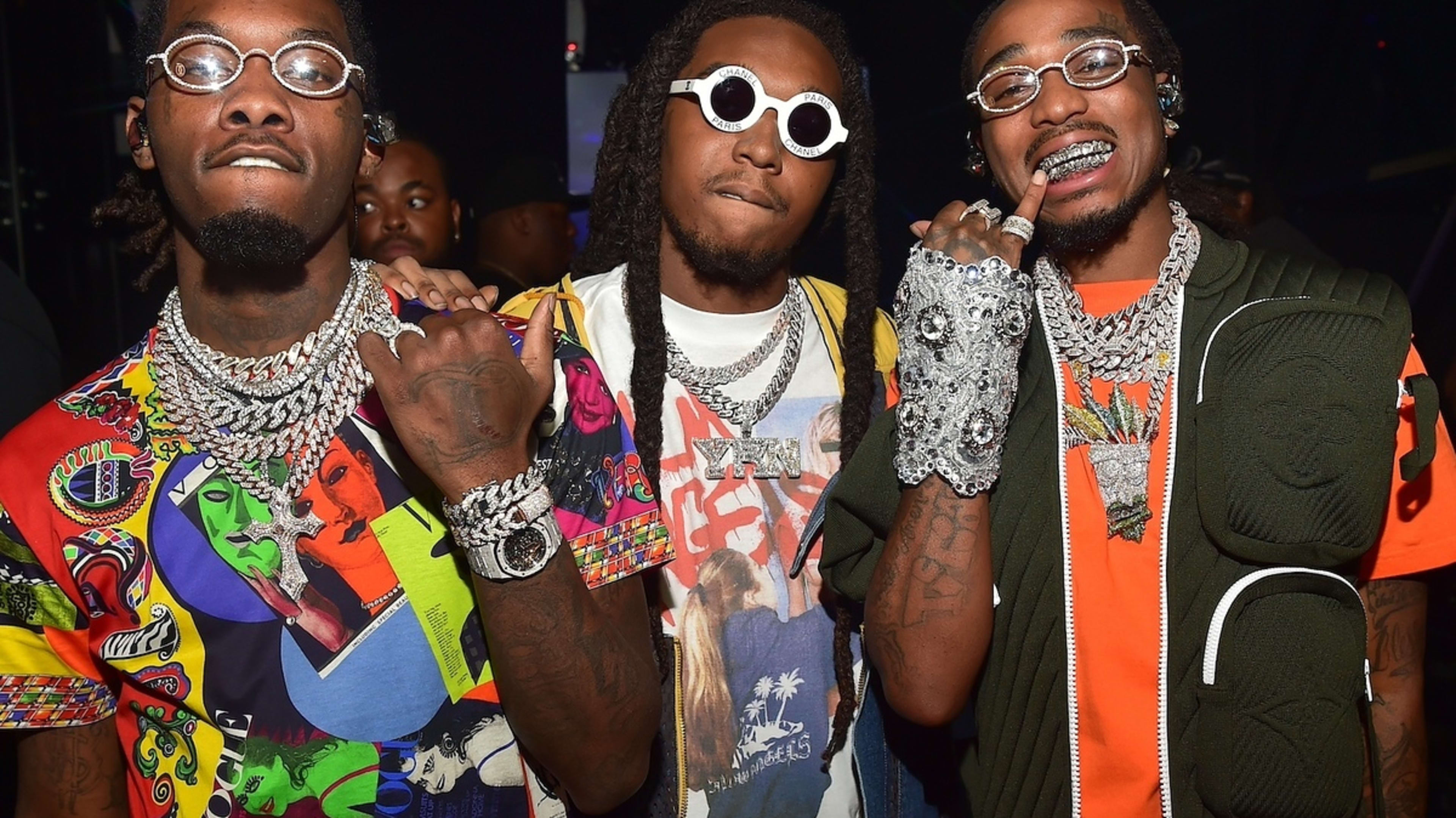 Three men posing together, two wearing glasses and abundant jewelry. The one in the middle has a graphic tee and vest