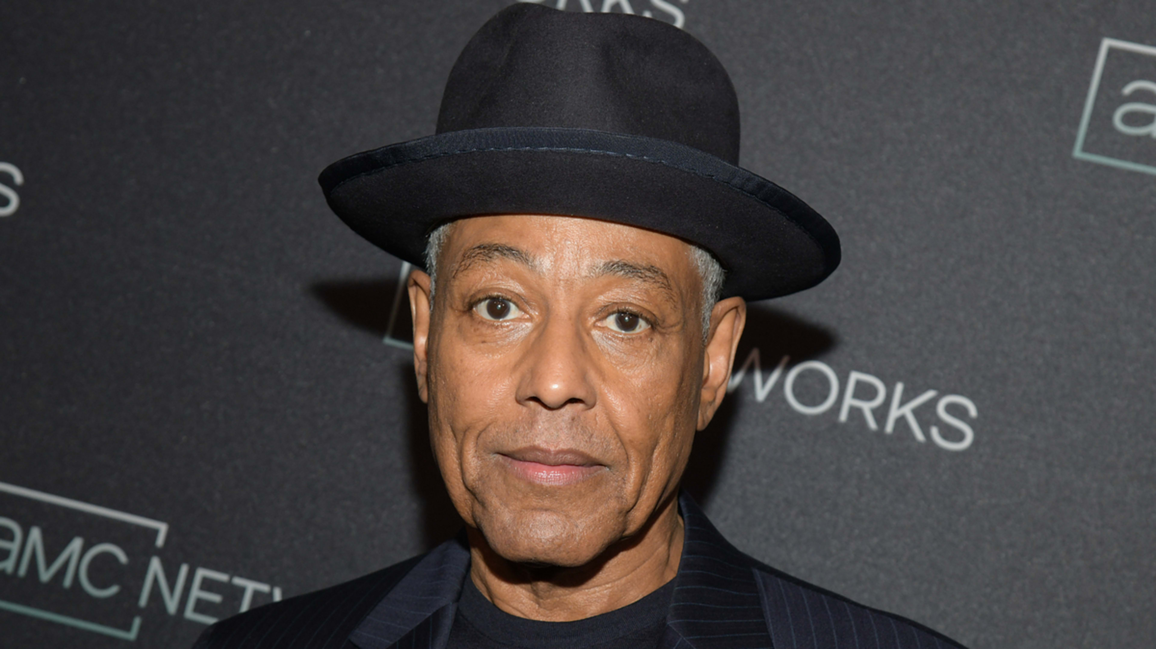 Giancarlo Esposito wearing a black suit and hat at an event with a logo backdrop