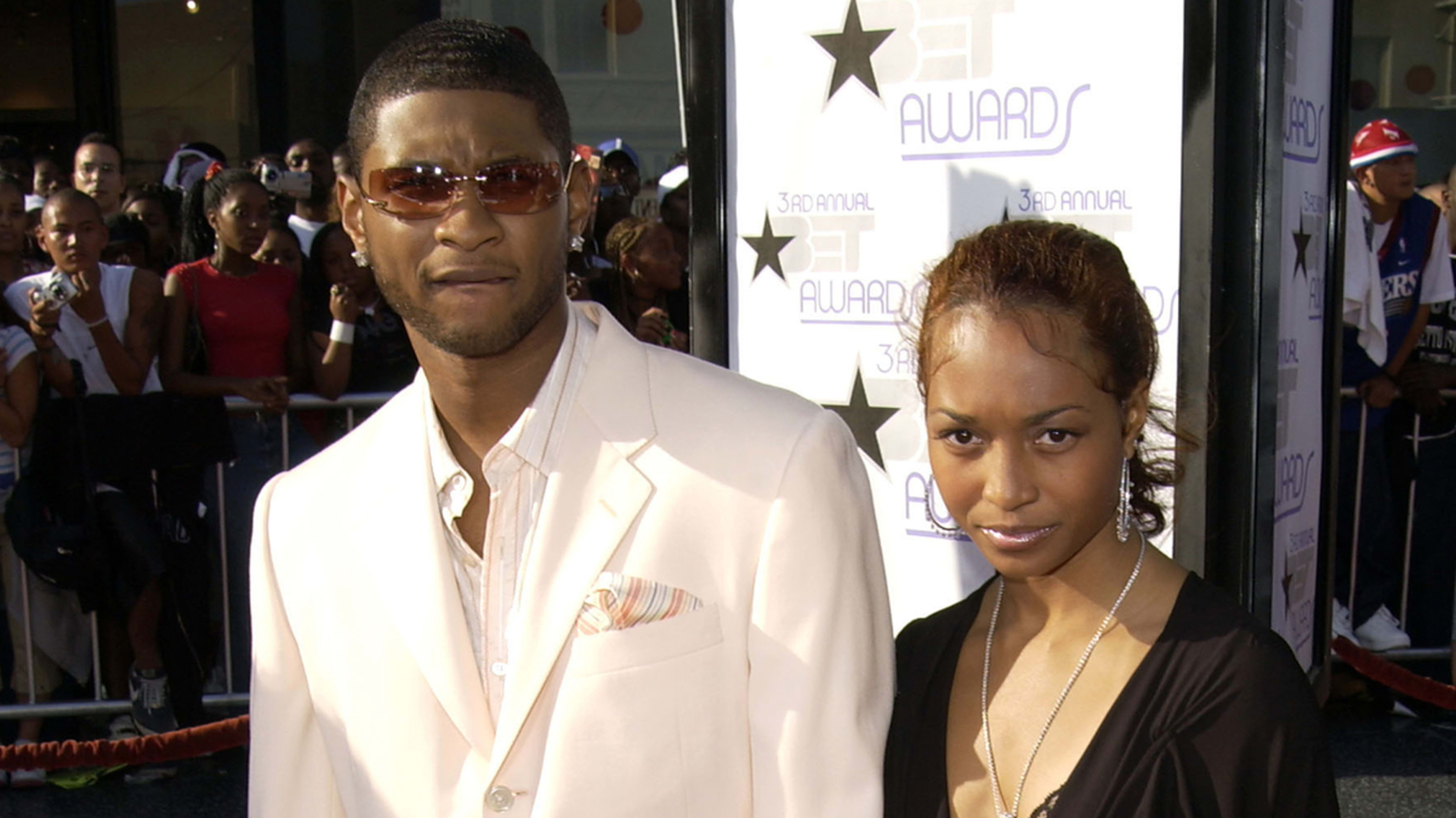 Two people posing at an event, one in a light suit and the other in a dark top and necklace