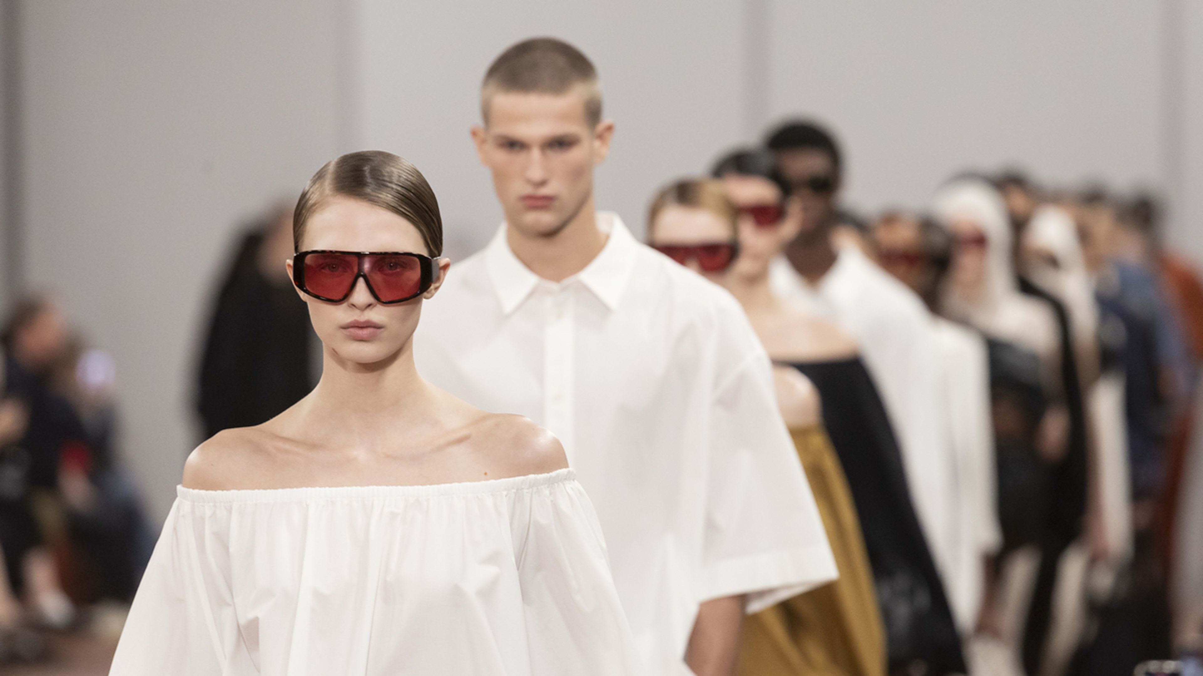 Models in a fashion show wearing minimalist outfits, with a focus on a female model in a white off-the-shoulder top