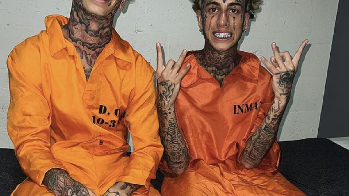 Two individuals, one with “D.G” on his chest, “10-3,” the other with "INM4TE", both heavily tattooed, wearing orange prison jumpsuits, sit on a bench making hand signs