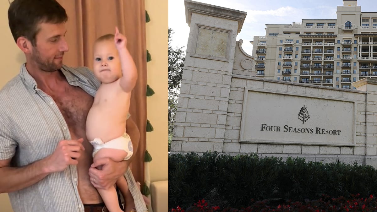 A man wearing a white shirt holds a baby in a diaper. Next to them, there is a sign that reads "Four Seasons Resort" in front of a large building