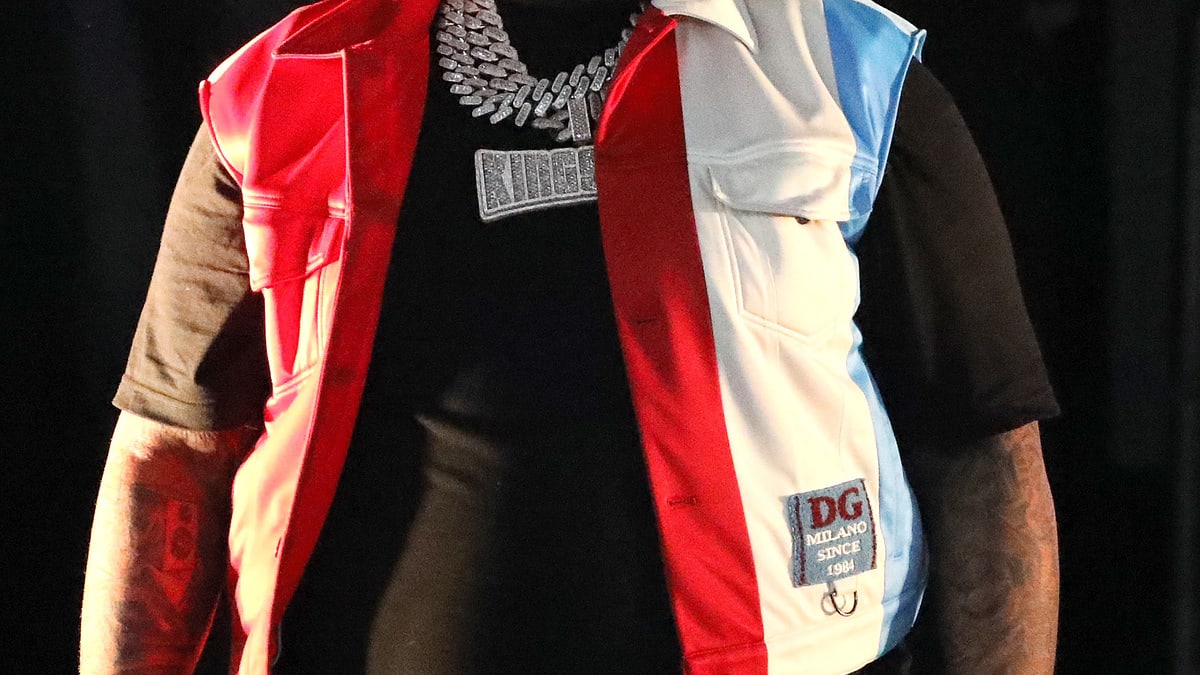 Sean Kingston wears a black outfit and a red, white, and blue vest, with heavy silver jewelry, while standing and holding a microphone