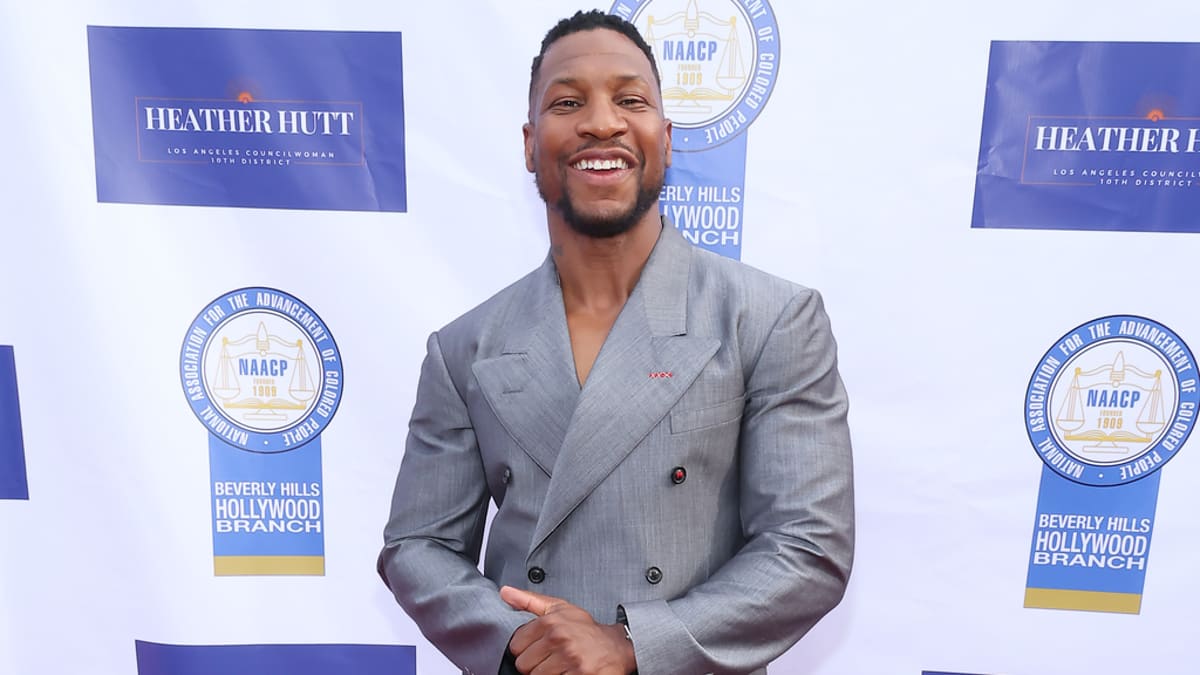 Jonathan Majors smiles on the red carpet in a gray double-breasted suit and black shoes at an event with NAACP and Beverly Hills Hollywood NAACP logos