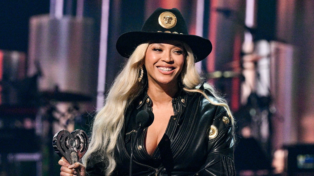 Beyoncé in a leather outfit and hat, holding an award at a ceremony, standing at a podium with a microphone, smiling
