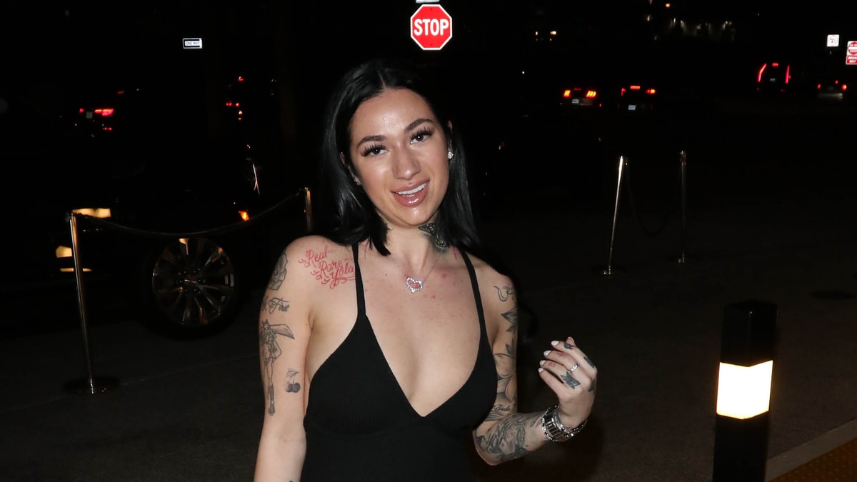 A woman with tattoos, dressed in a black sleeveless top, poses outdoors at night near a stop sign