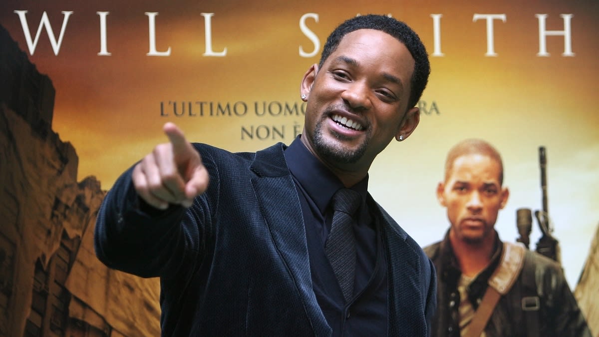 Will Smith in a dark suit smiling and pointing, standing in front of a poster featuring his name and an image of himself from a movie