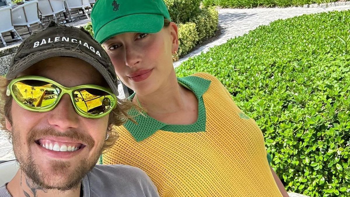 Justin Bieber, in casual wear and sunglasses, poses with Hailey Bieber, who wears a yellow top and green hat, holding her baby bump. Outdoor background with greenery