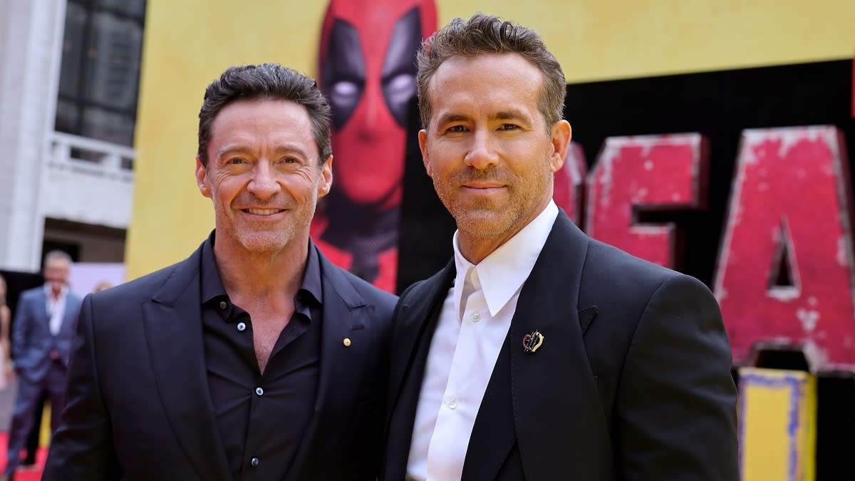 Hugh Jackman in a black suit and Ryan Reynolds in a black blazer over a white shirt, standing together at a movie premiere with Deadpool artwork in the background
