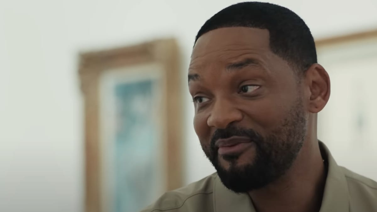 Will Smith, wearing a casual open-neck shirt, smiles warmly in this close-up shot. The photo is likely taken indoors with framed artwork visible in the background