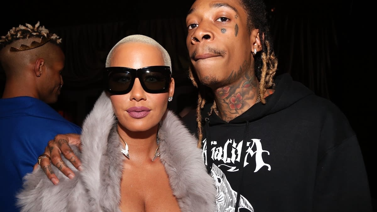 Amber Rose wearing large sunglasses and a fur-trimmed coat, stands beside Wiz Khalifa, who wears a hoodie with tattoos visible on his face