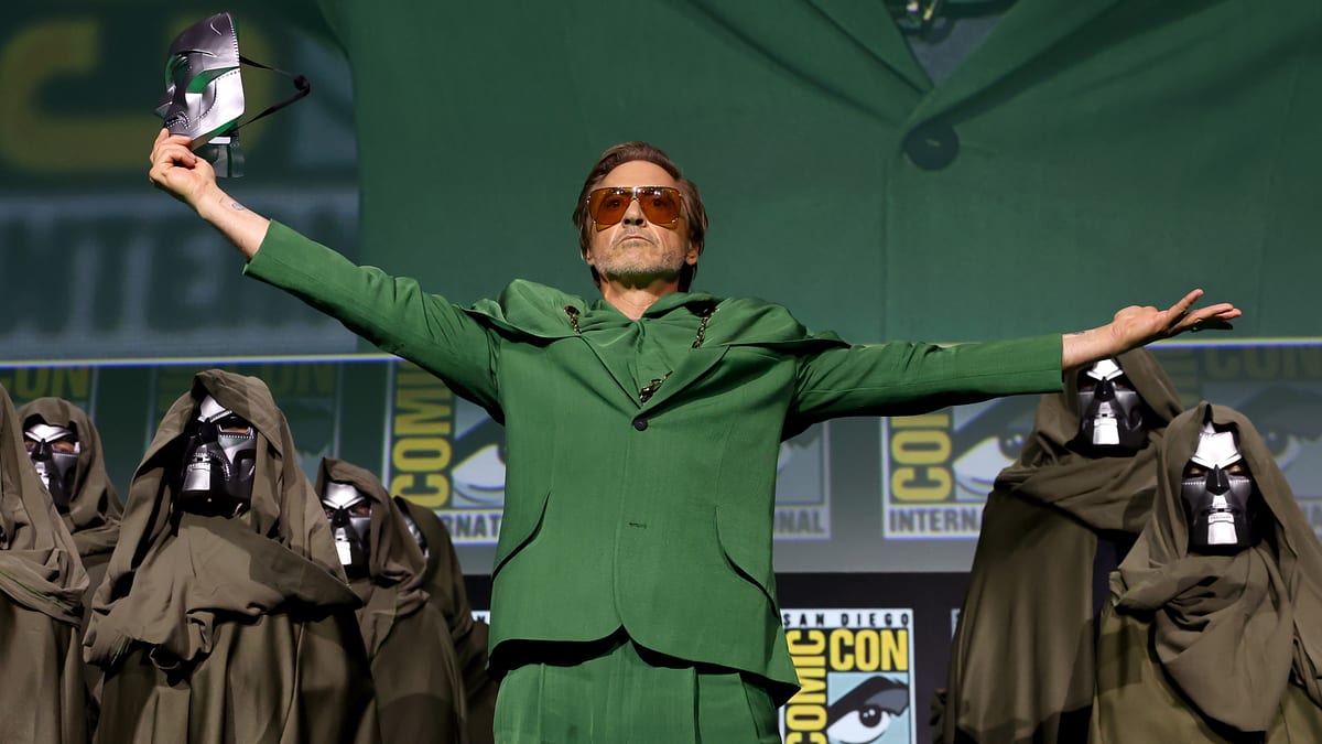 Robert Downey Jr. on stage at Comic-Con, wearing a green suit with arms outstretched, surrounded by people in hooded cloaks and metallic masks