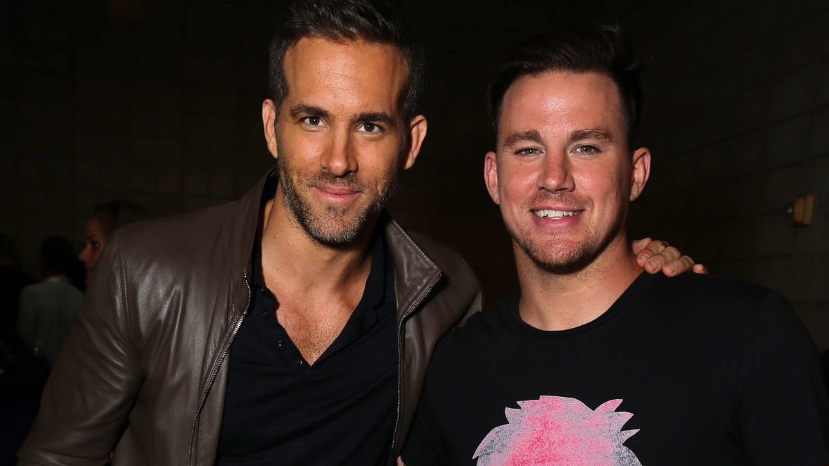 Ryan Reynolds, in a leather jacket and dark jeans, stands with Channing Tatum, in a graphic tee and jeans, both smiling at the camera