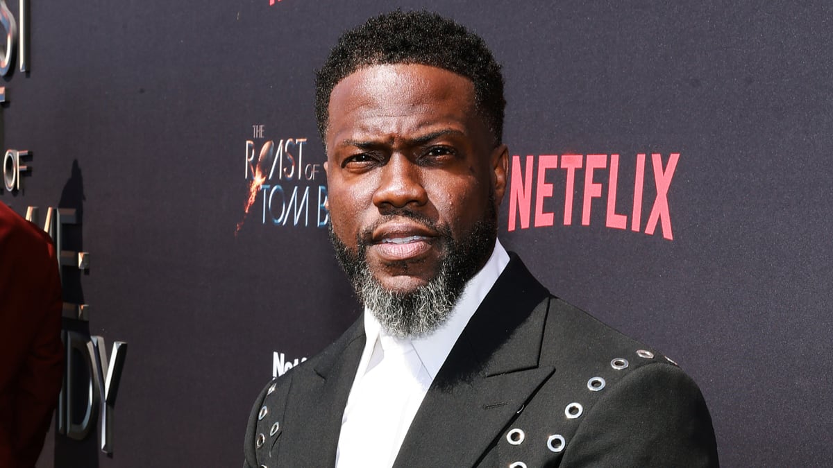 Kevin Hart on the red carpet, wearing a black jacket with metallic grommet details, white shirt, neatly trimmed beard and short hair. Netflix logo in background