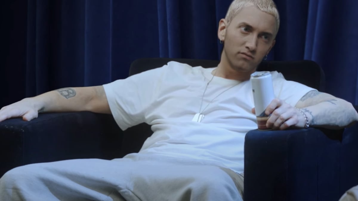 Eminem sits on a black chair, wearing a white shirt and grey pants, holding a beverage can, against a blue curtain backdrop
