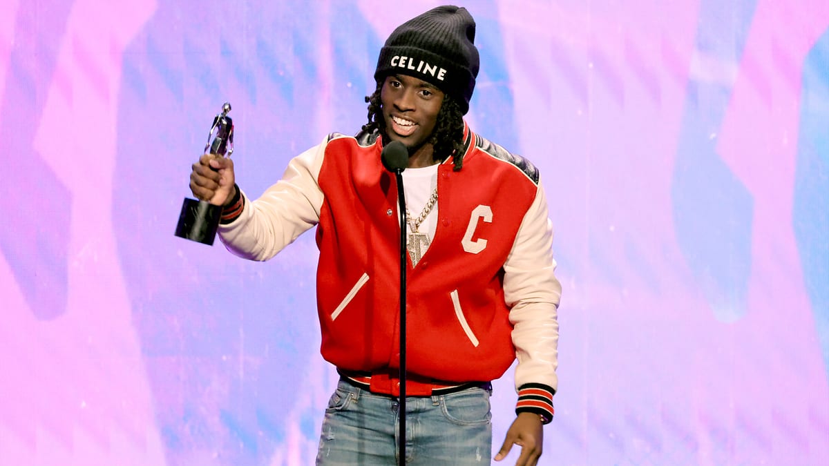 Kai Cenat holds an award while speaking into a microphone. He is wearing a red varsity jacket, black beanie, jeans, and multiple gold chains