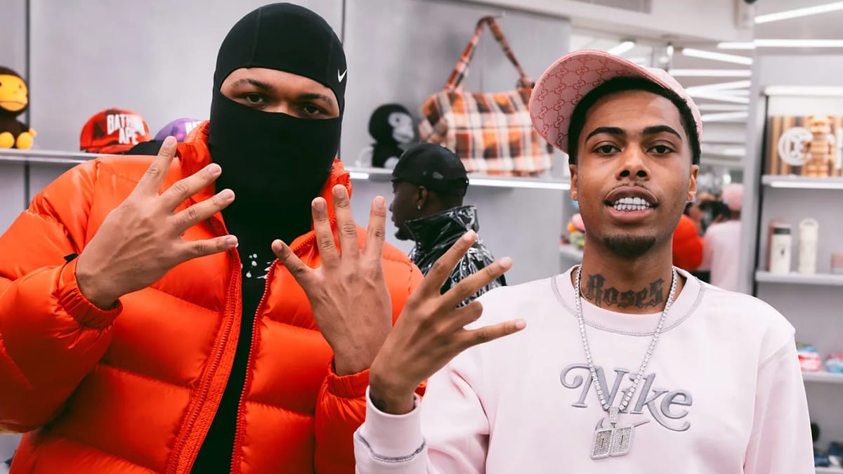 Unknown persons in a clothing store, one in a mask and orange jacket, the other in a pink Nike sweater and hat, displaying hand gestures