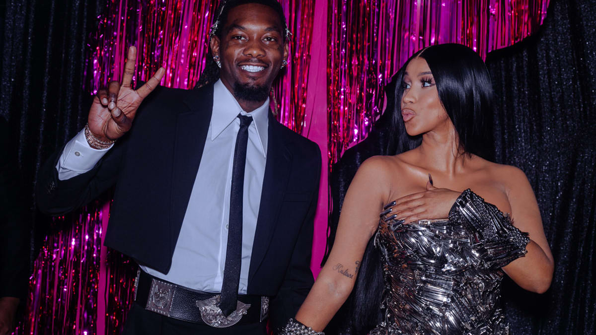 Offset and Cardi B pose in stylish outfits at an event with shiny decorative background. Offset wears a black suit while Cardi B wears a metallic dress