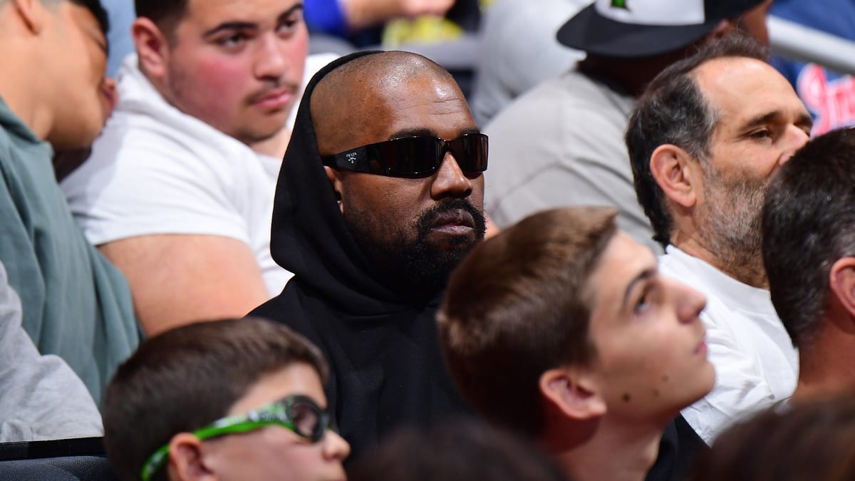 Kanye West in the audience at a music event, wearing dark sunglasses and a hoodie, surrounded by other attendees