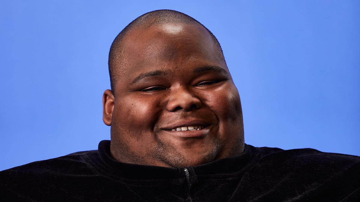 A smiling person with a shaved head and a velvet top, against a plain blue background