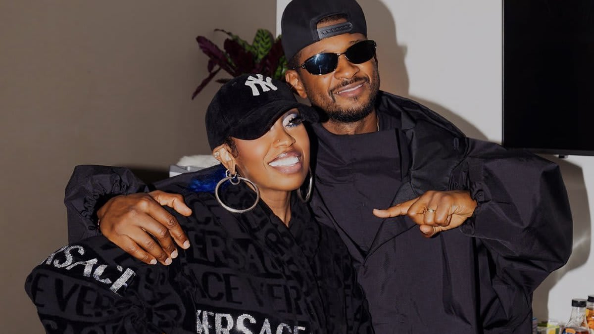 Missy Elliott and Usher are smiling and posing together. Missy wears a Versace robe and cap, holding a bouquet. Usher is in a black jacket and sunglasses