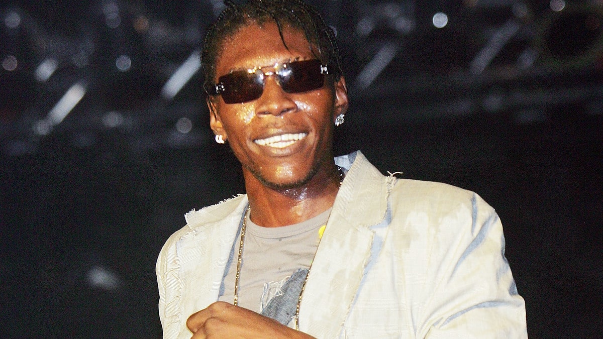 Vybz Kartel, on stage, smiling in sunglasses, a light jacket over a printed t-shirt, and jeans, holding a microphone