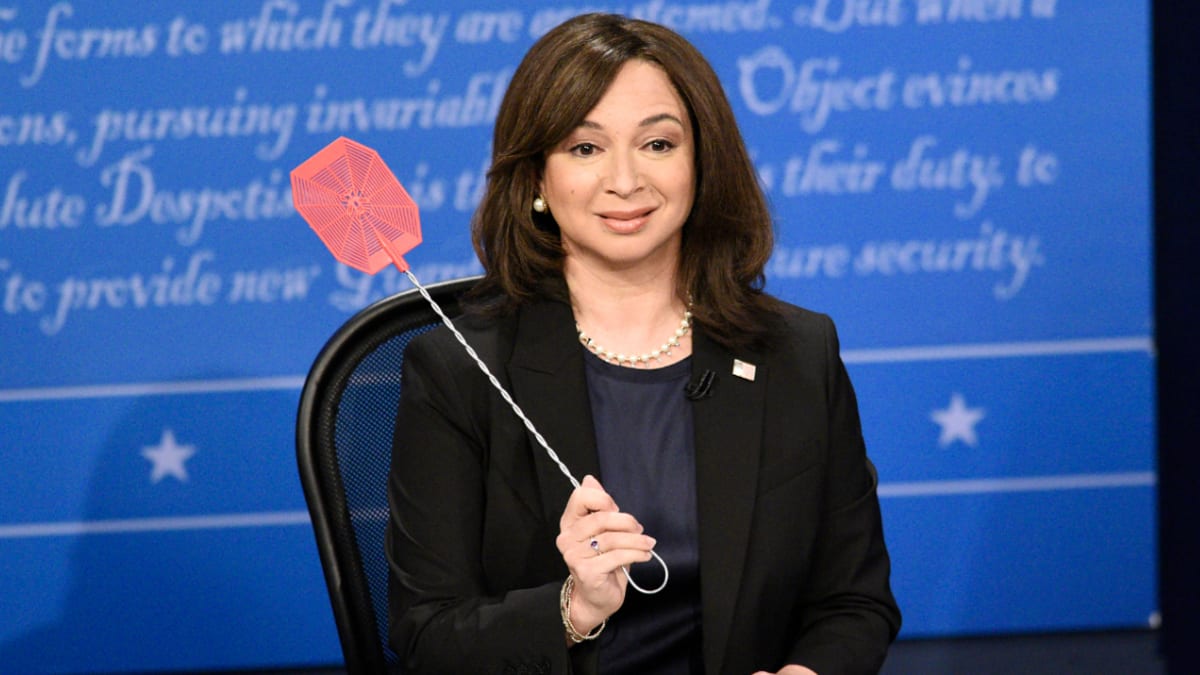 Maya Rudolph, dressed in formal attire as Kamala Harris, holds a prop fly swatter during a comedic skit on a blue-themed stage with text in the background