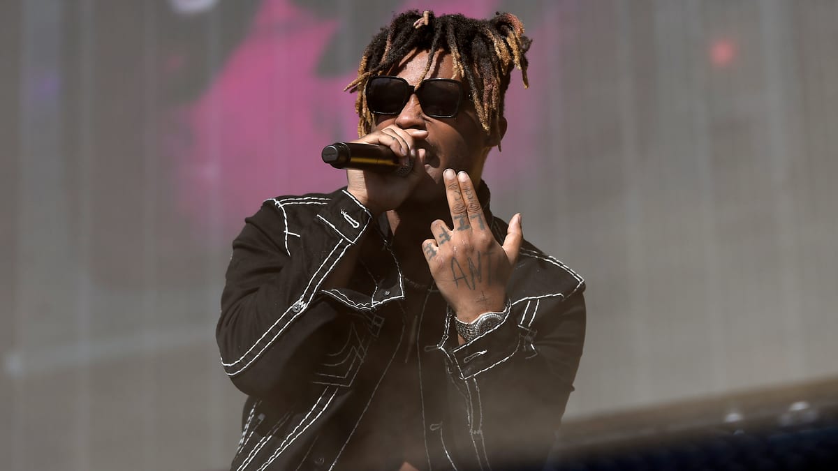 Juice WRLD performing on stage, wearing a black jacket with white stitching, sunglasses, and holding a microphone