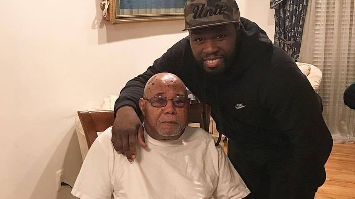 50 Cent, wearing a black Nike hoodie and a cap that says "Unit," smiles and poses with an elderly man seated in a chair at home