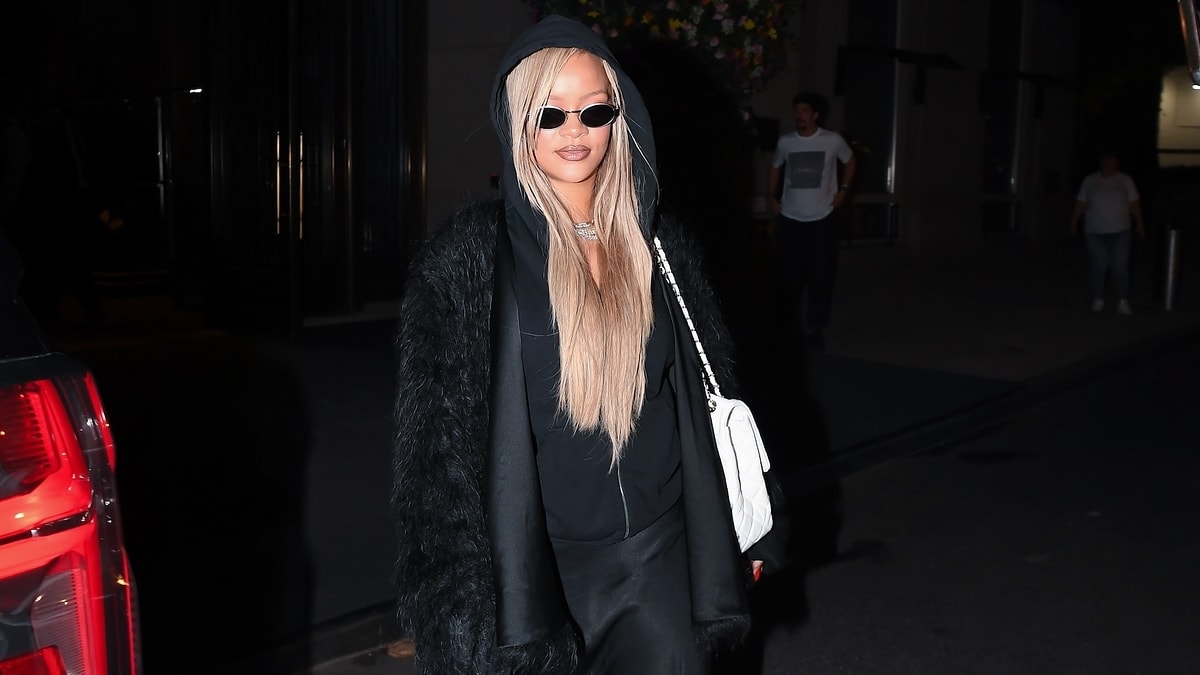 Rita Ora wearing a black outfit with a hood and long blonde hair, holding a white handbag, walking at night